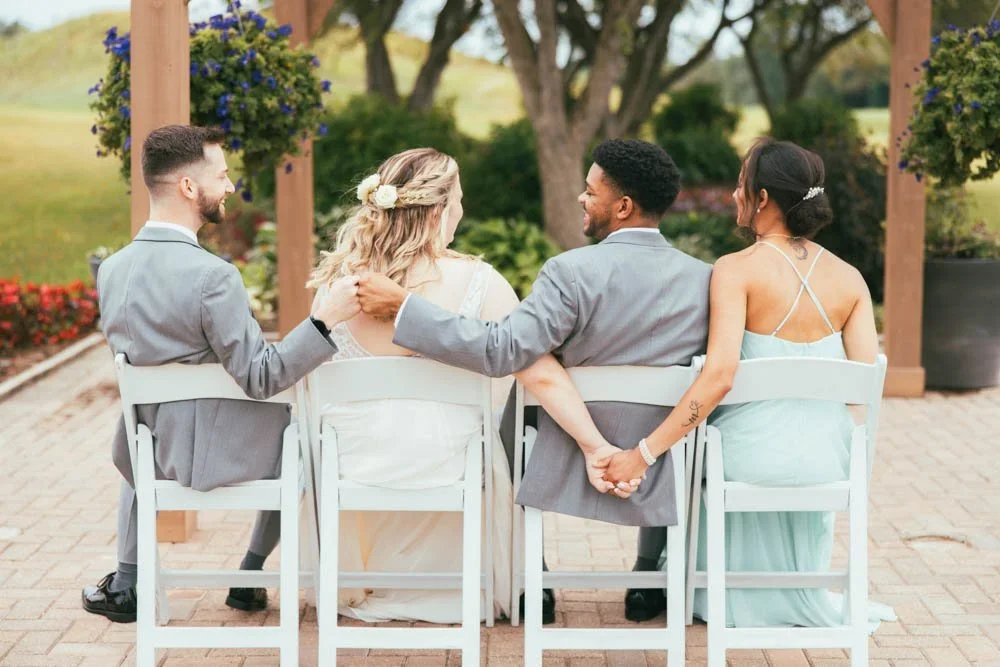 A group of four people, two men and two women, sitting on white chairs outdoors during a wedding or formal event. They are seated with their backs to the camera, holding hands, and engaging in conversation. The setting has trees, colorful flowers, and a wooden pergola with hanging flower baskets.