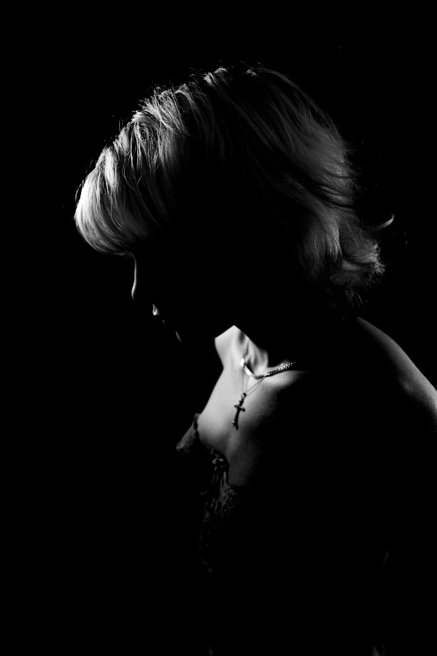 Silhouette of a woman with short hair wearing a necklace with a cross pendant, photographed in black and white against a dark background.