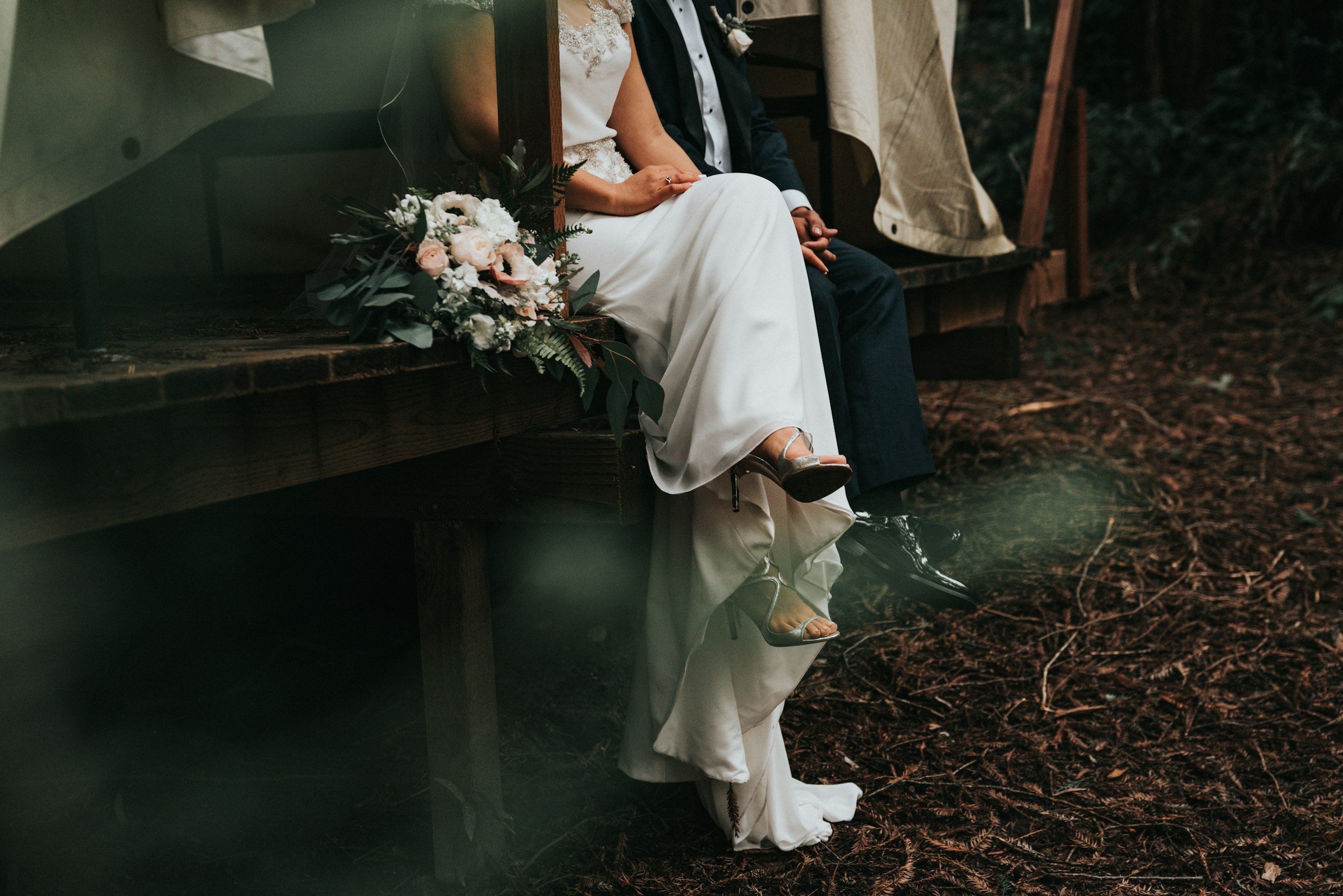 Details of a bride and groom sitting on a wooden platform amid a forest, with a bouquet of flowers beside them.