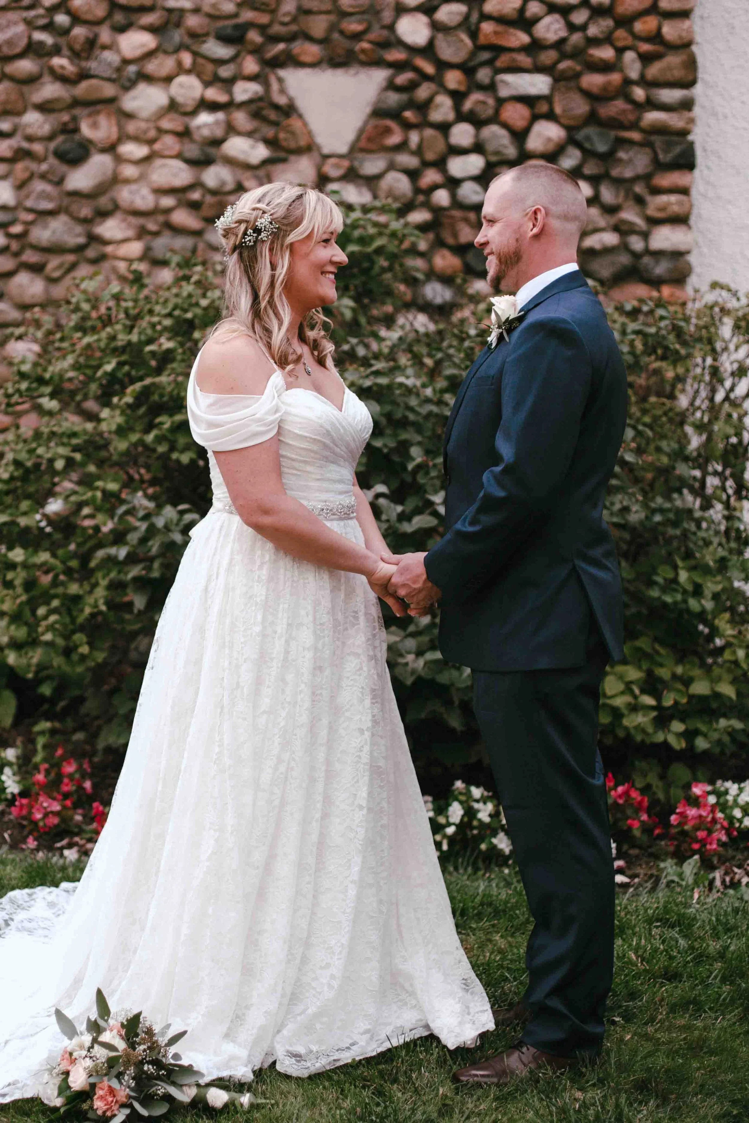 A bride and groom holding hands and smiling at each other during their wedding ceremony outdoors, with a stone wall and greenery in the background.