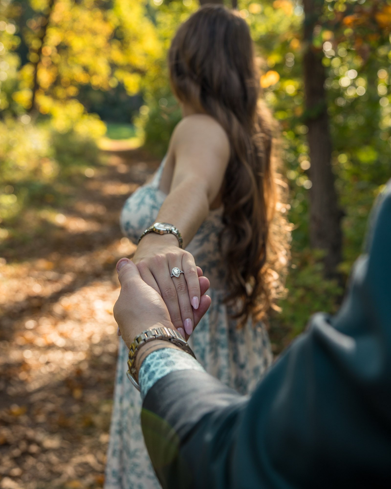 Ann Arbor engagement photography of A woman with long brown hair standing in a wooded area, holding hands with a person in a suit, showcasing an engagement ring on her finger.