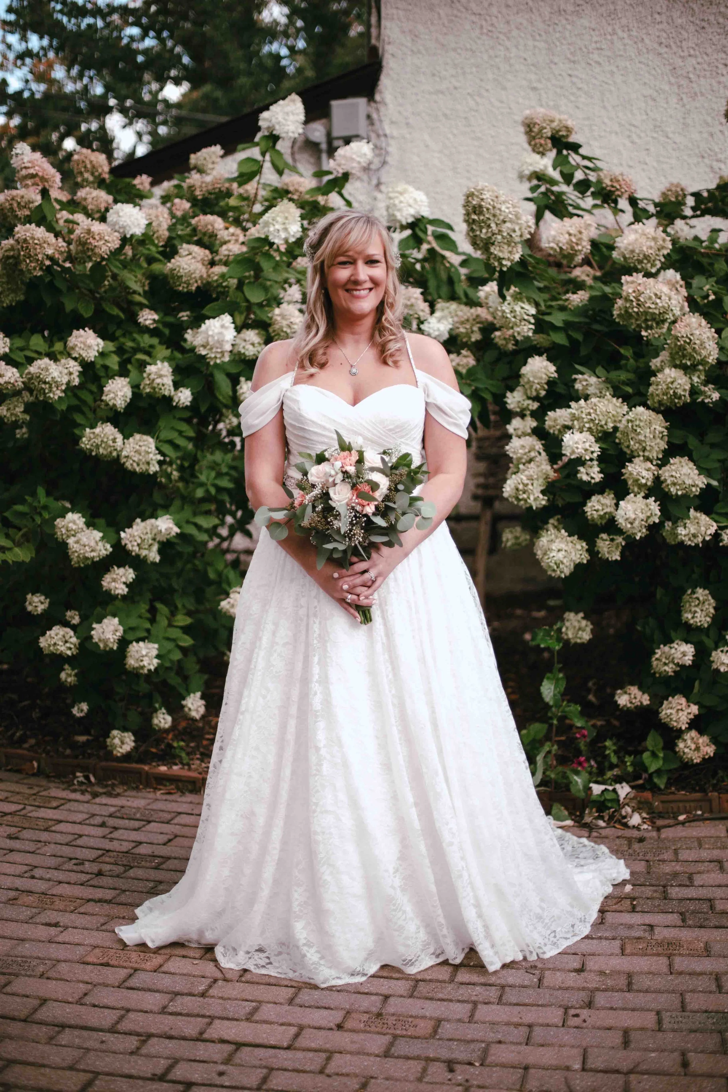 A woman in a white wedding dress holding a bouquet, standing on a brick path with a large bush of white flowers behind her, smiling.