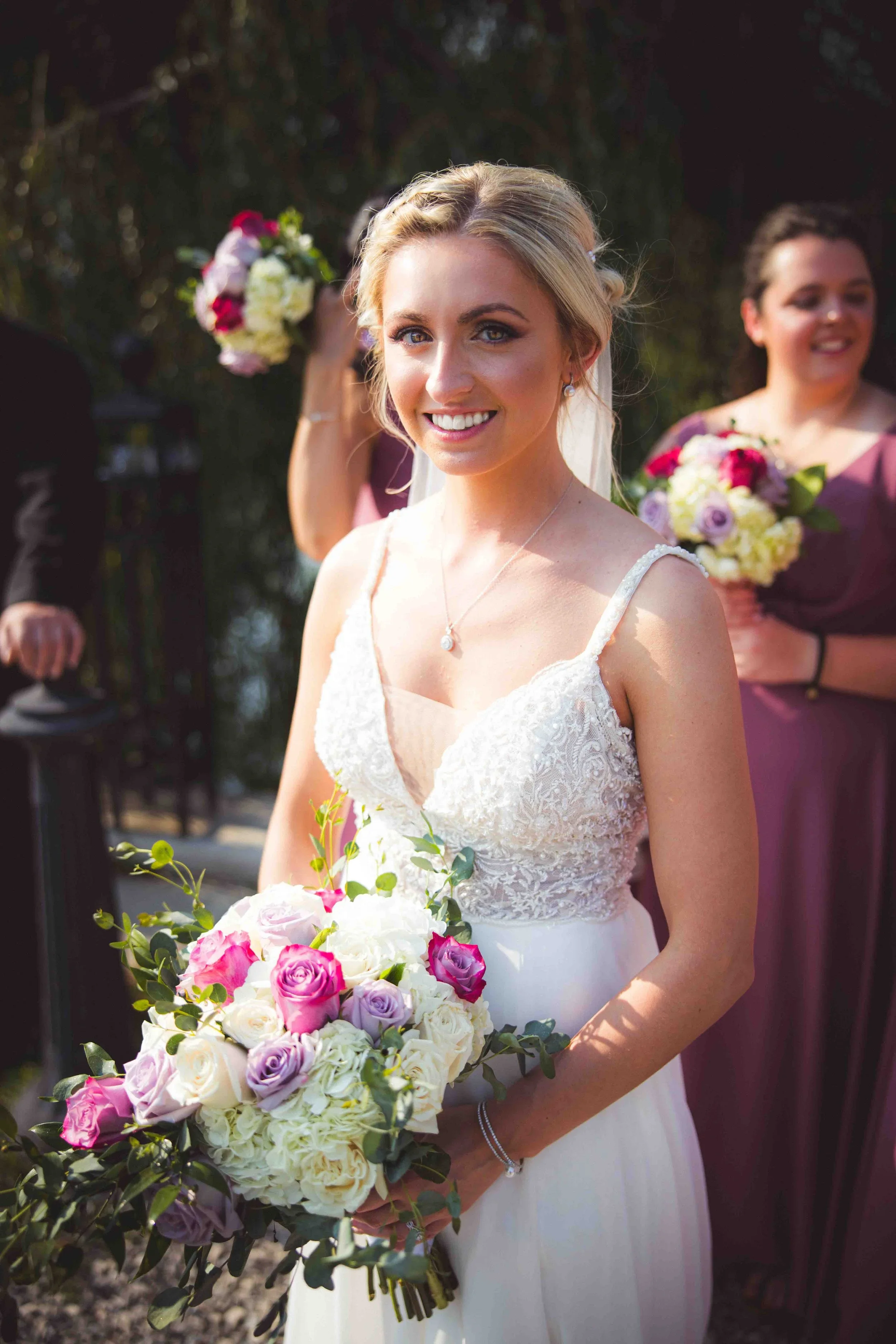 A smiling bride holding a bouquet of pink, purple, and white flowers during a wedding ceremony.