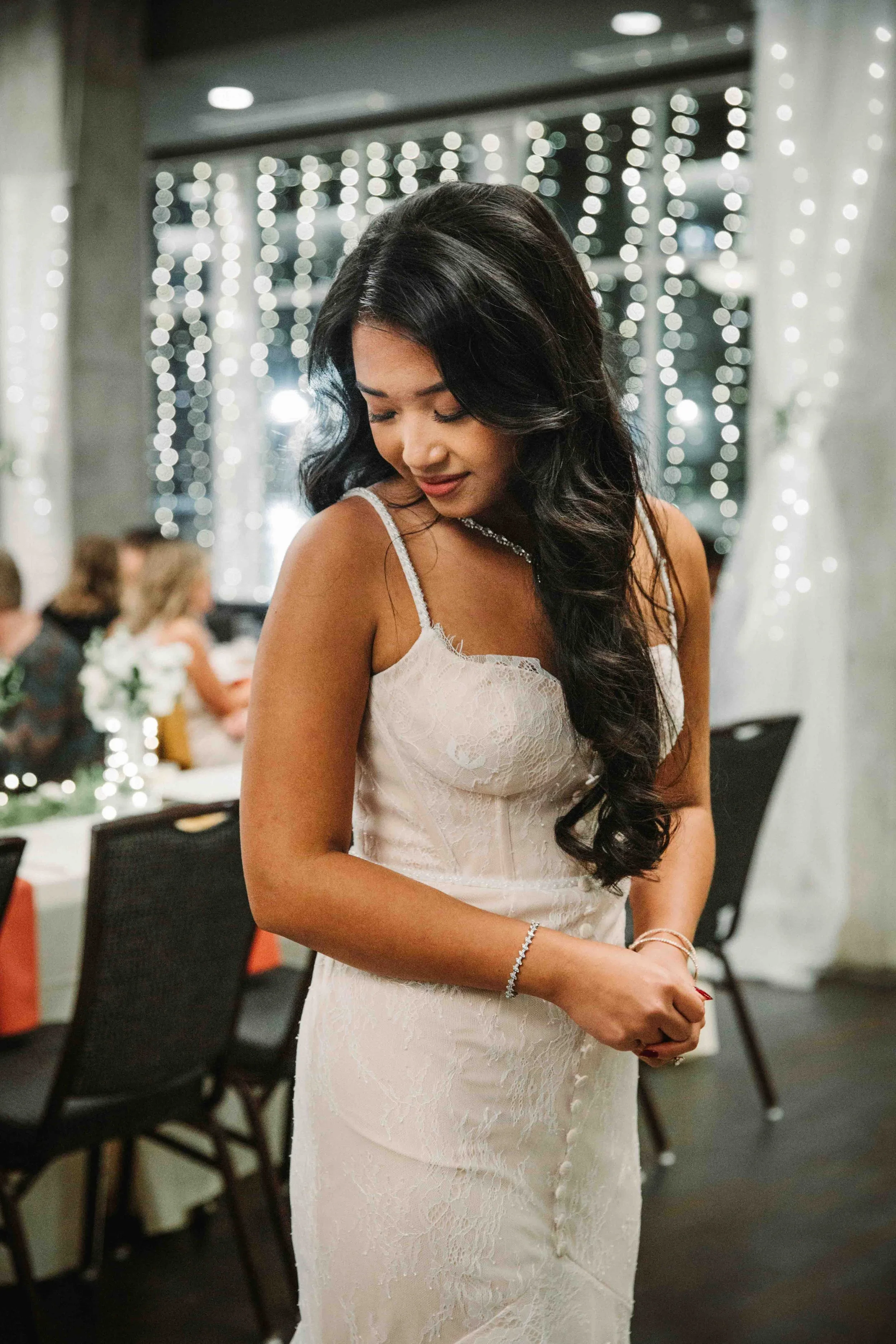 A woman in a white lace dress with thin straps, standing at an event with a decorated background of string lights and guests at tables, looking down with a gentle expression.