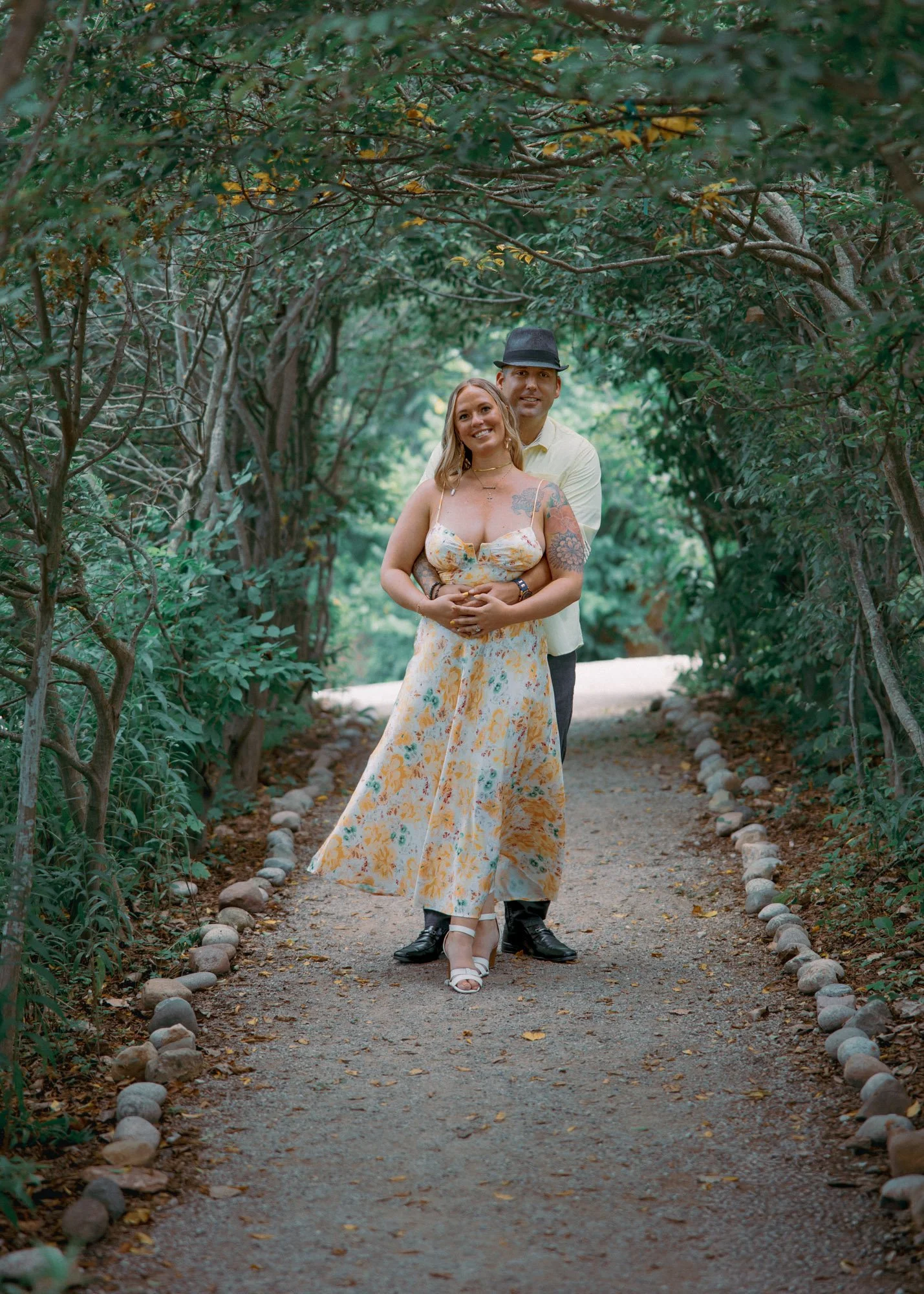 A couple standing on a gravel path framed by green trees, embracing each other and smiling at the camera