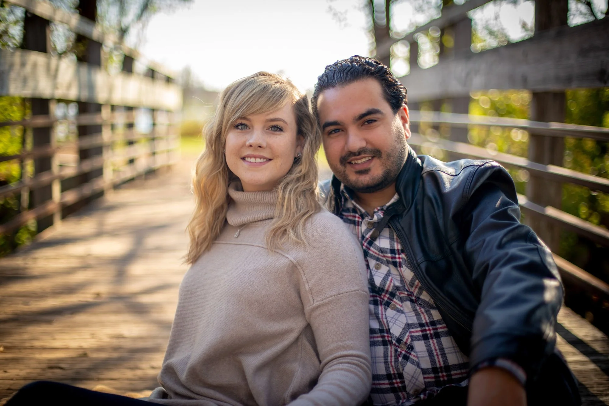 A young woman with blonde hair and a young man with dark hair sitting closely together on a wooden bridge outdoors, smiling at the camera.