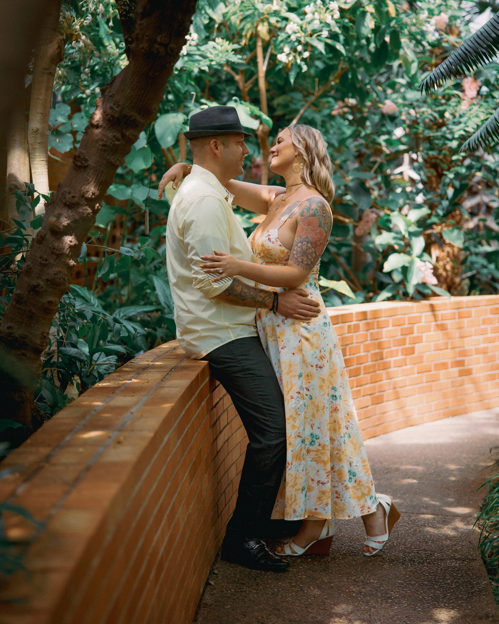 A couple with tattoos sharing a romantic moment in a lush, green garden, sitting on a brick ledge, with the woman dressed in a floral dress and the man in a light-colored shirt and black hat.