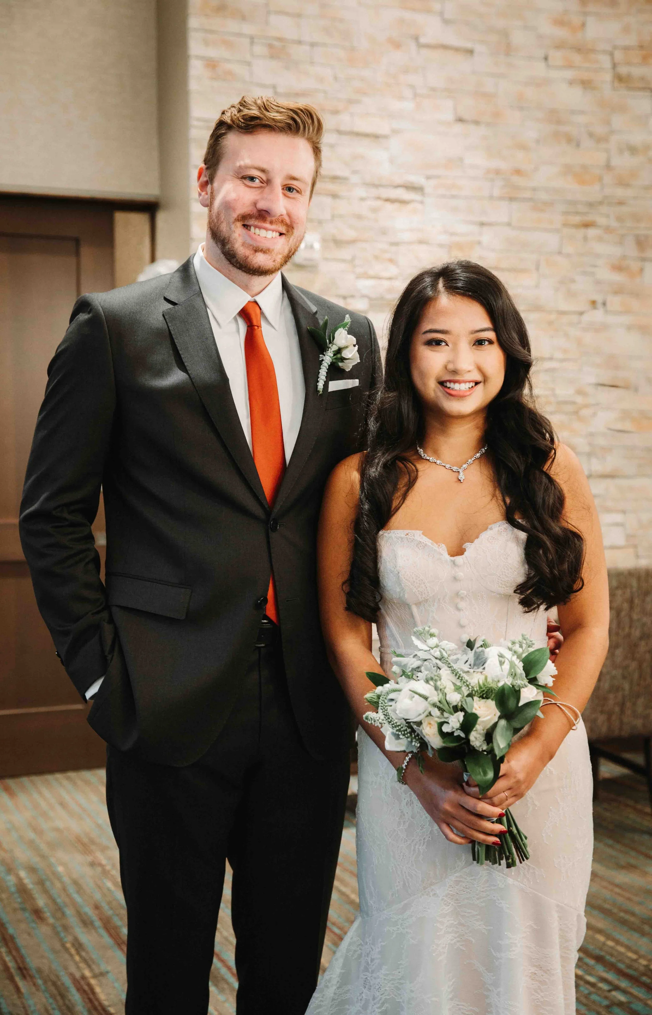 A smiling couple dressed in wedding attire posing indoors against a brick wall. The man wears a dark suit with a white shirt, orange tie, and boutonniere. The woman wears a white lace wedding dress with a necklace and holds a bouquet of white flowers and greenery.