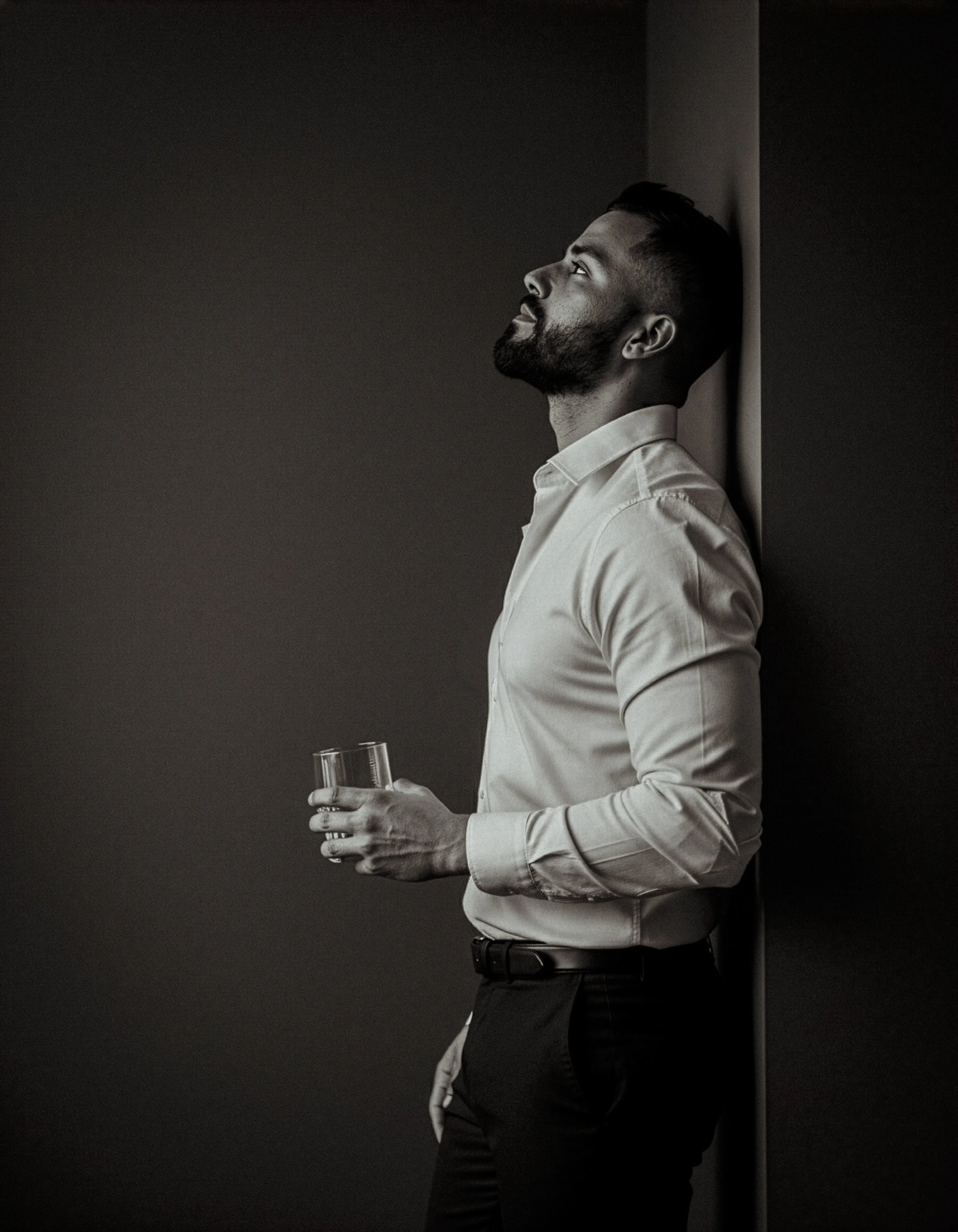 A man with a beard and short hair, dressed in a light-colored button-up shirt and dark pants, stands against a dark wall holding a glass of water, looking upward with a thoughtful expression.