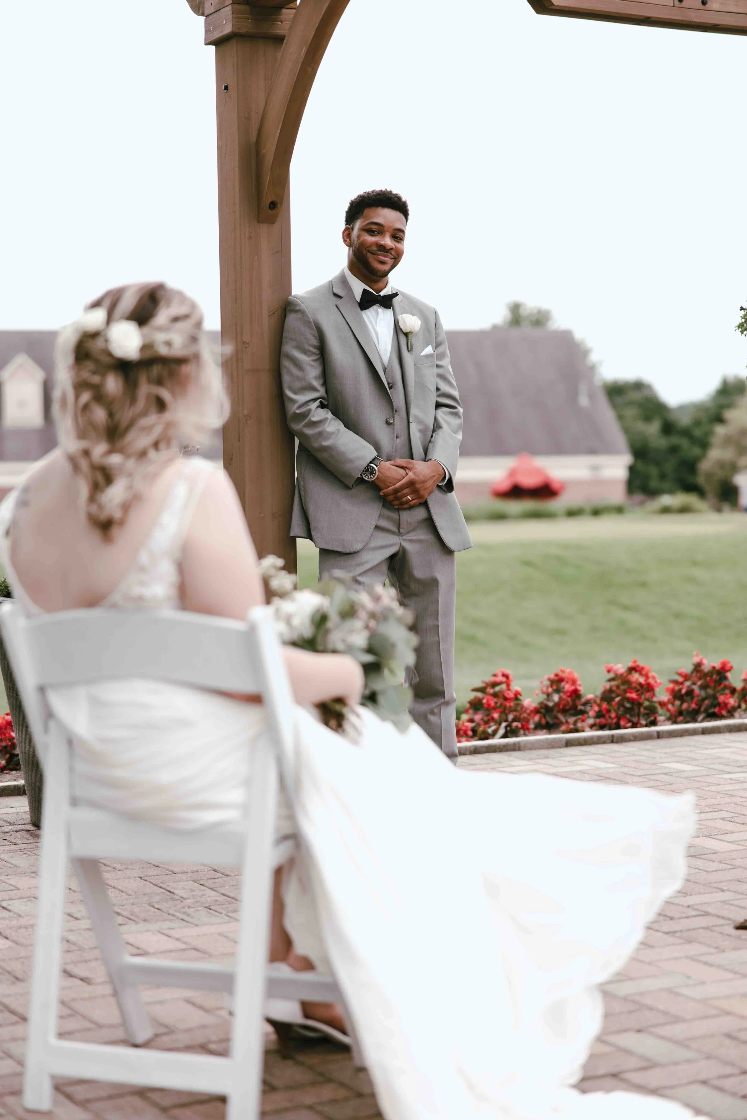 Ypsilanti wedding photography of A groom standing outdoors in a gray suit, black bow tie, and white shirt, smiling and looking at a seated bride with blonde hair, wearing a white wedding dress, holding a bouquet, at a wedding ceremony.