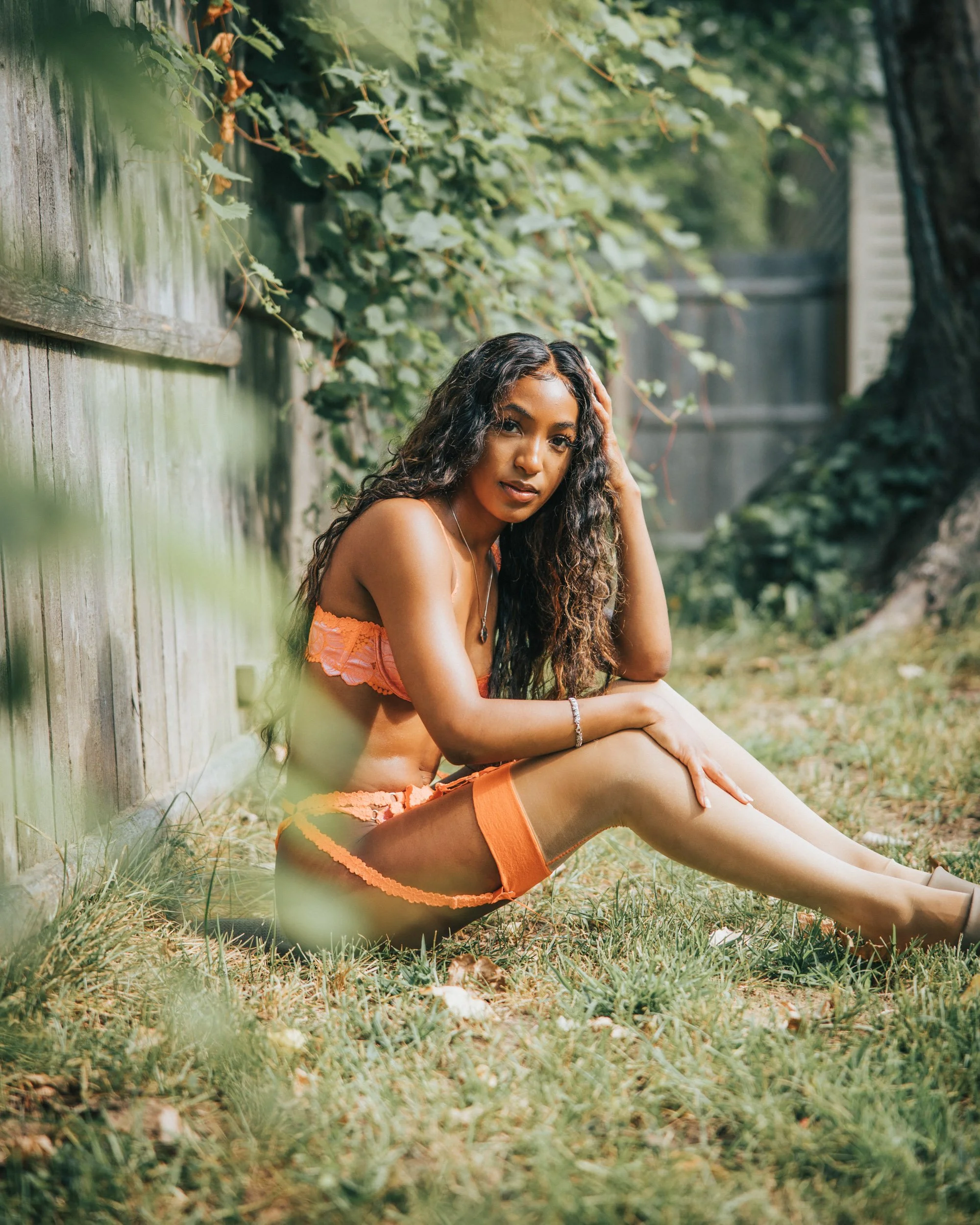 A woman with long curly hair sitting on grass beside a weathered wooden fence, wearing orange lingerie and thigh-high stockings, surrounded by greenery.