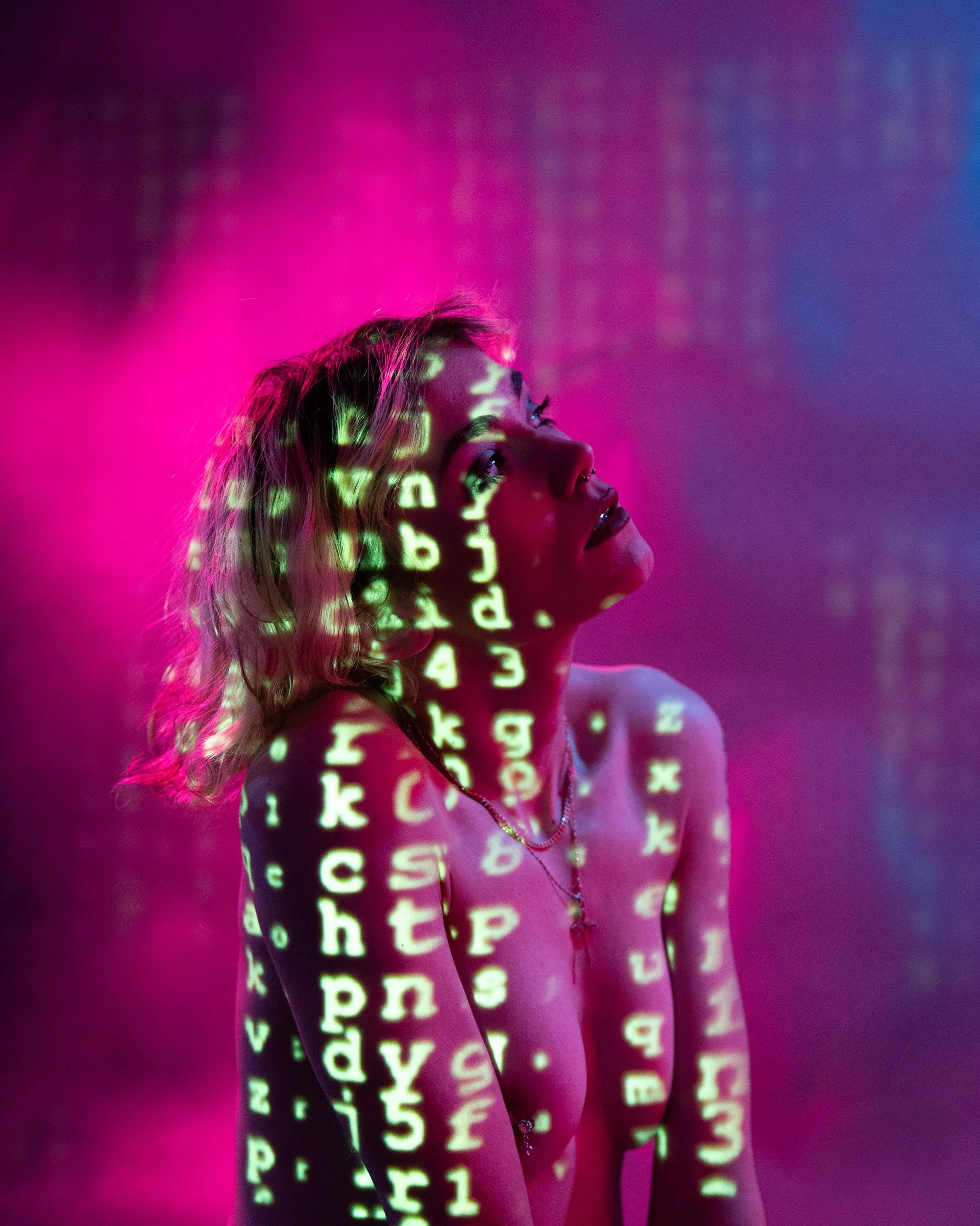 A woman with blonde hair is illuminated by pink and purple neon light, with green computer code projected onto her face, body, and background.