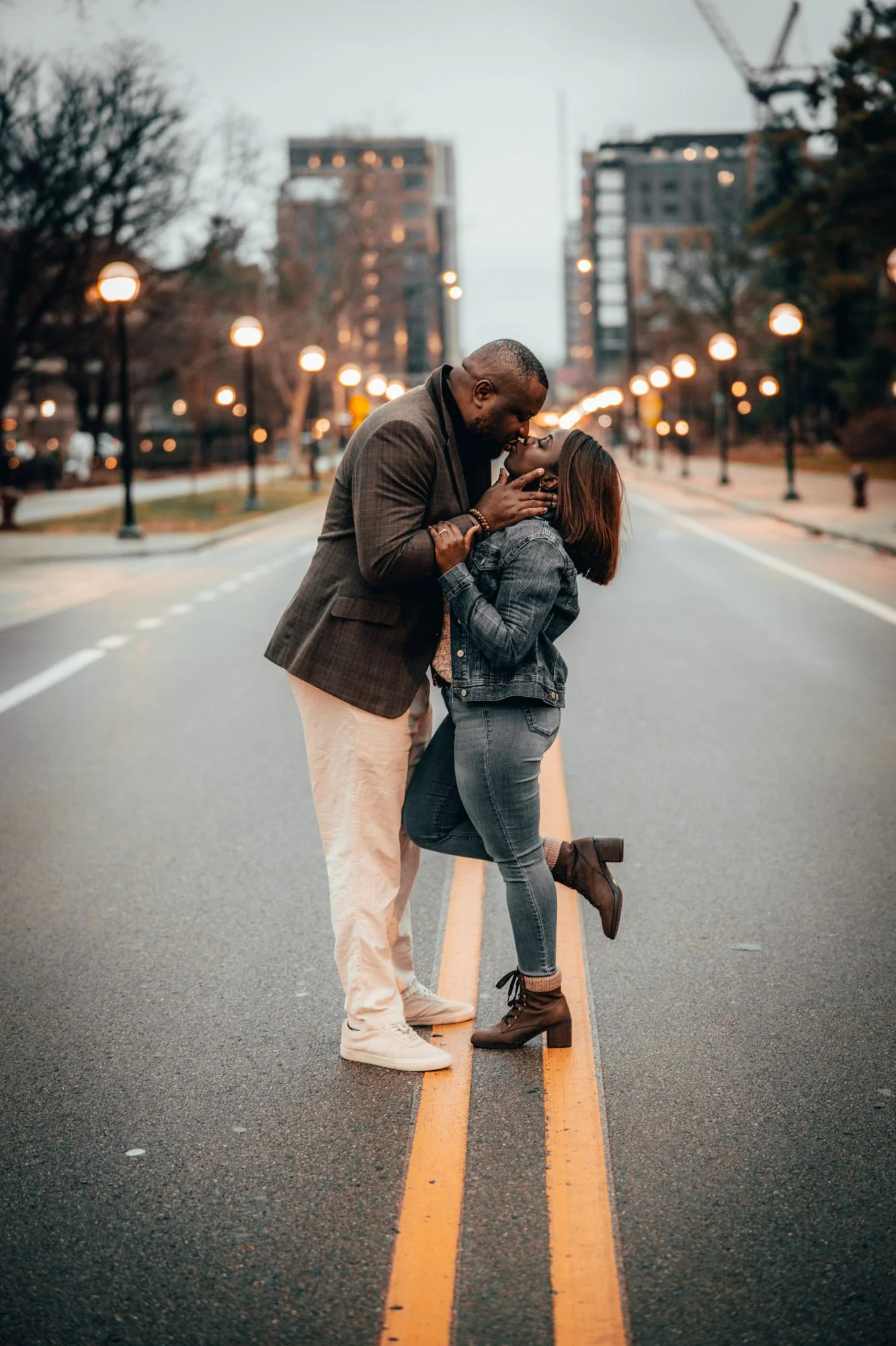 Ypsilanti Michigan engagement photography of A couple kissing on a city street at dusk, with streetlights and buildings visible in the background.