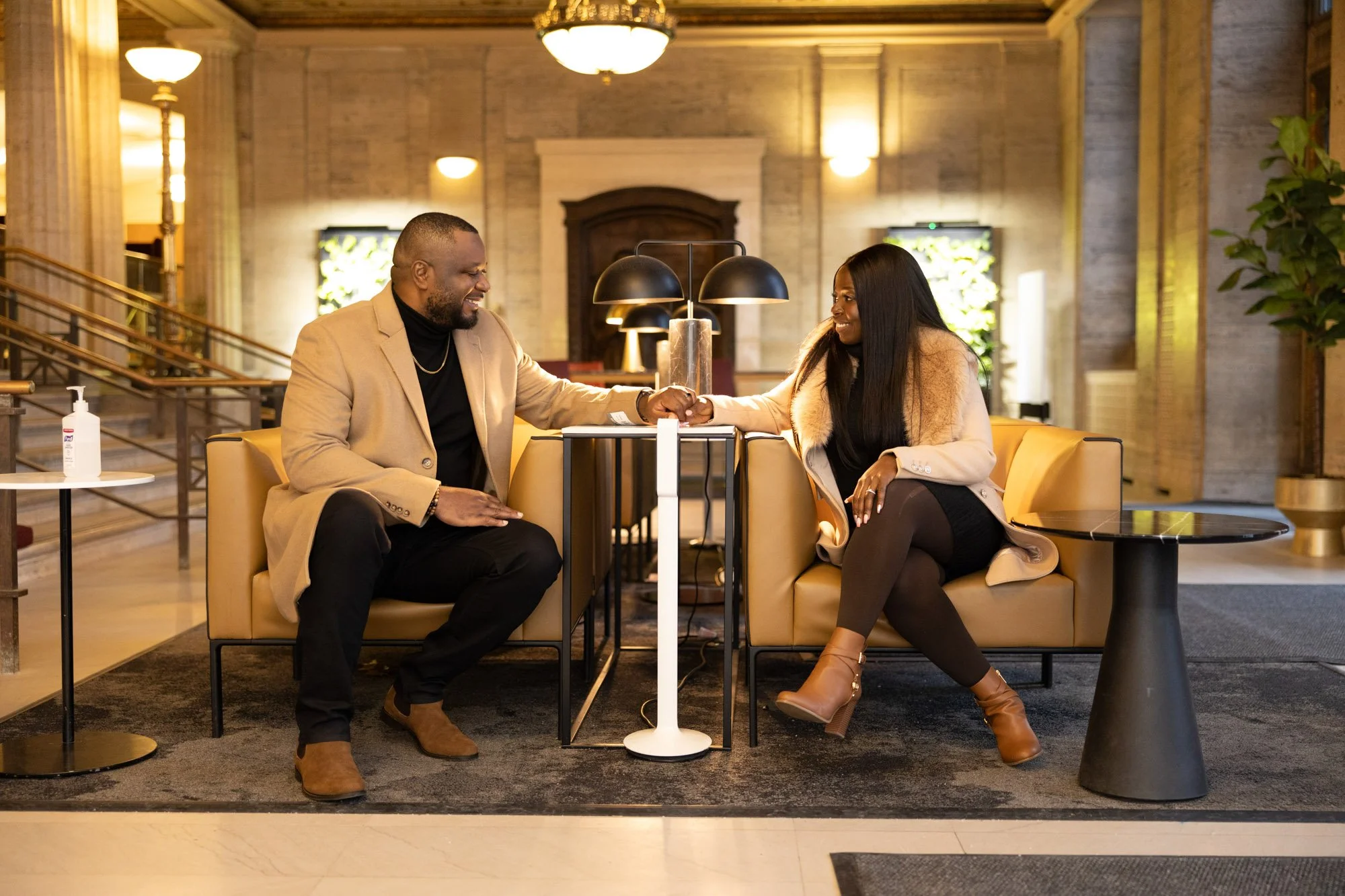 A man and a woman sitting on a yellow couch in a warmly lit hotel lobby, holding hands and smiling at each other.