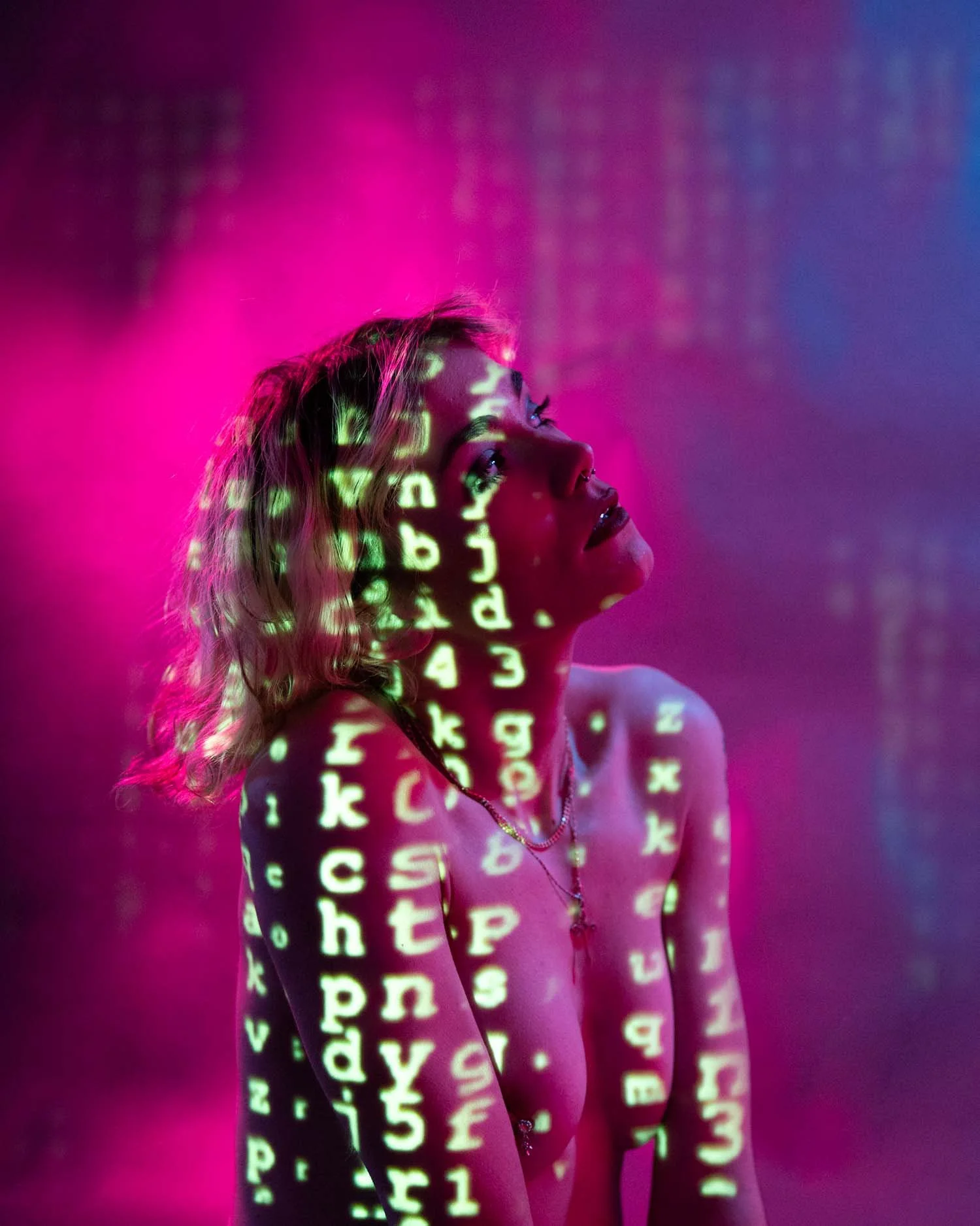 A woman with blonde hair looking up, with green and pink computer code projected onto her face and body, in a pink-lit environment