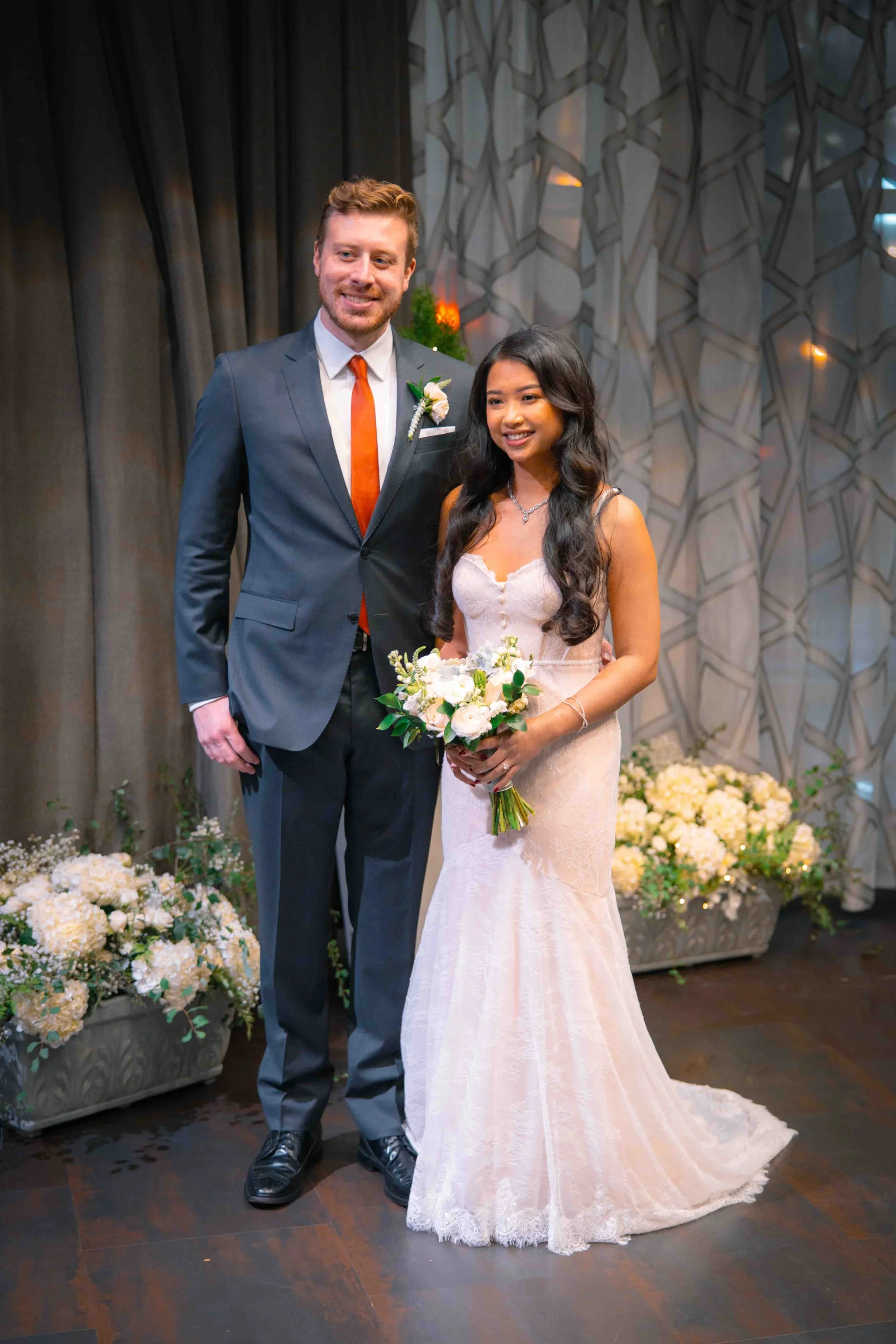 A bride in a white wedding gown holding a bouquet and a groom in a dark suit with a white shirt and orange tie, standing together at a wedding reception with floral decorations.