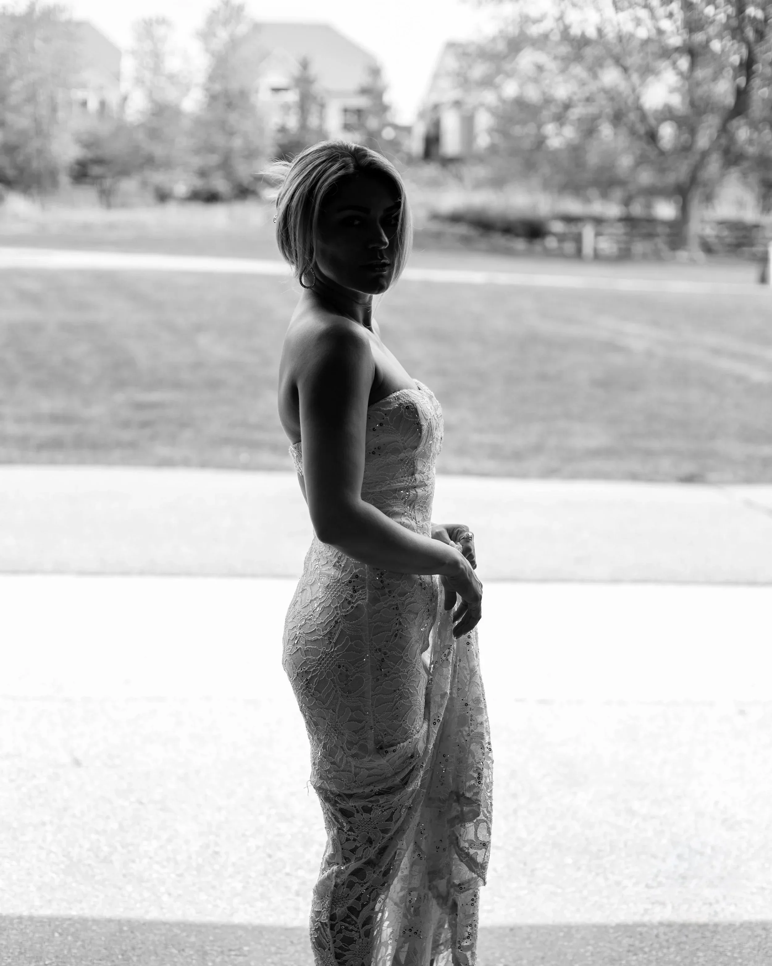 A woman in a strapless lace dress stands in front of a window, with a blurred outdoor scene in the background.