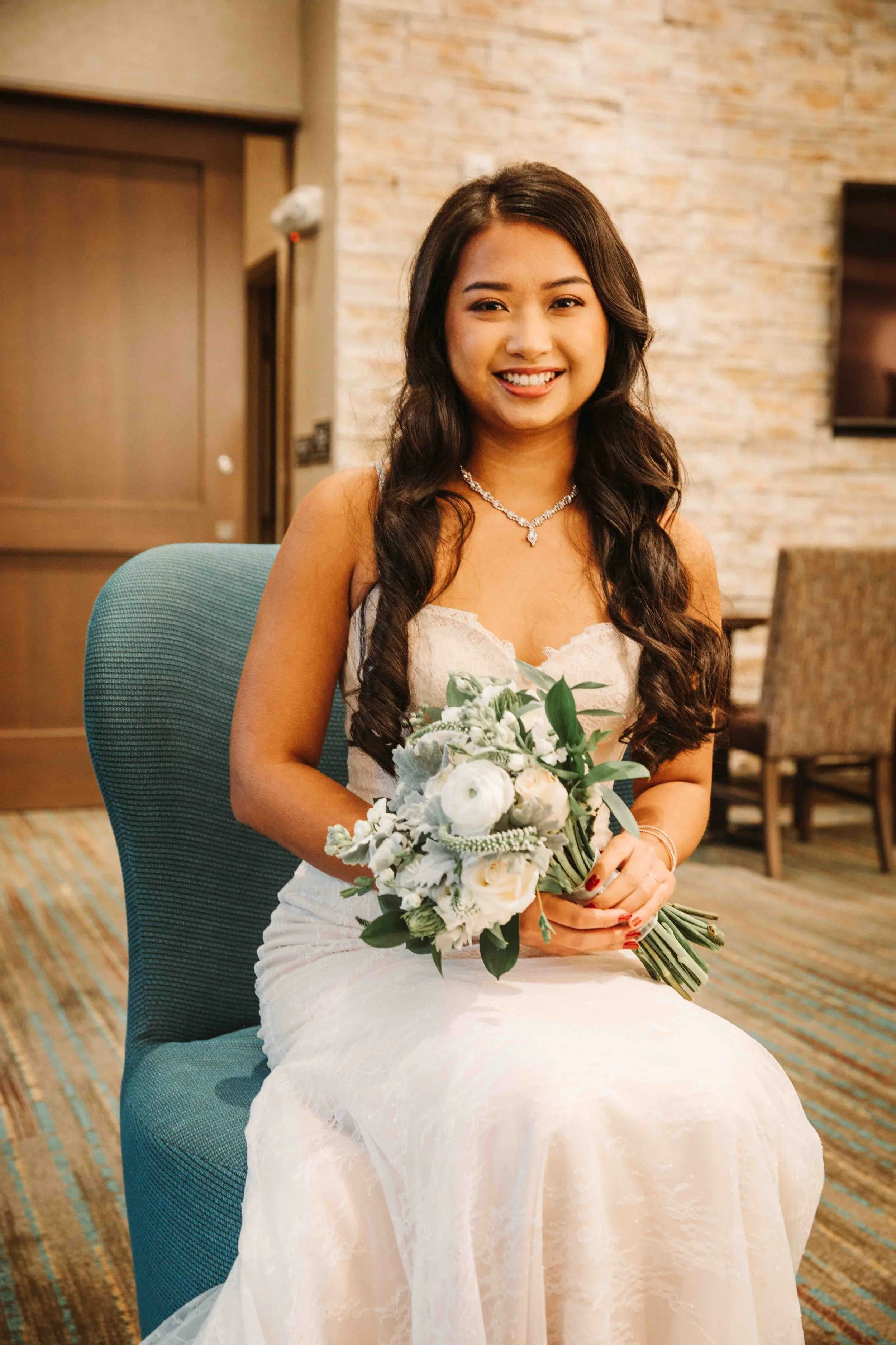 A smiling woman in a wedding dress holding a bouquet of white flowers, sitting on a teal chair indoors with a background of wood paneling and chairs.