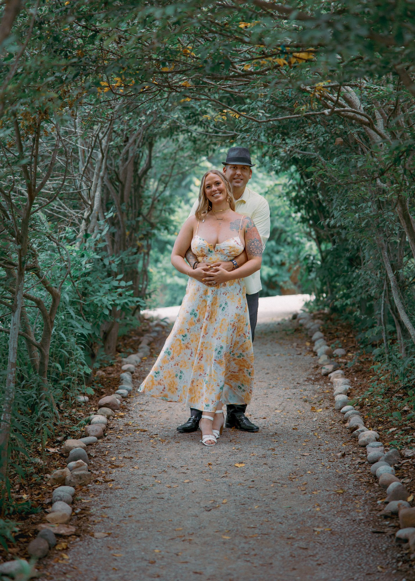 A couple standing on a wooded pathway, smiling. The woman is wearing a floral dress and white heels, and the man is dressed in a light shirt, dark pants, and a hat, wrapping her in his arms.
