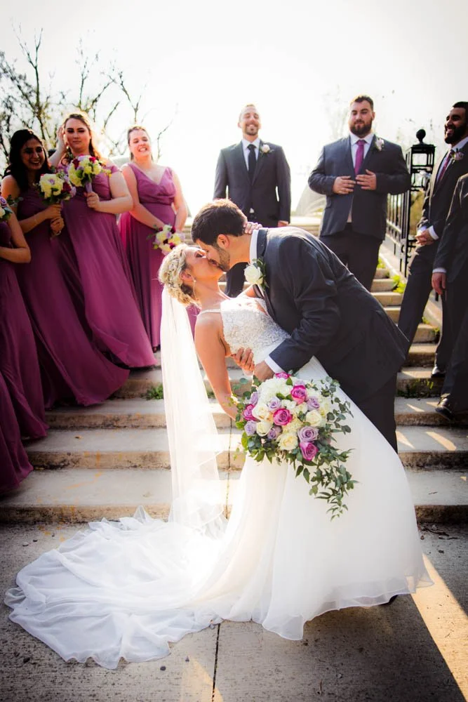 Ann Arbor Wedding photography of aBride and groom share a kiss on outdoor steps during wedding ceremony, surrounded by bridesmaids in purple dresses and groomsmen in suits, with sunlight in the background.