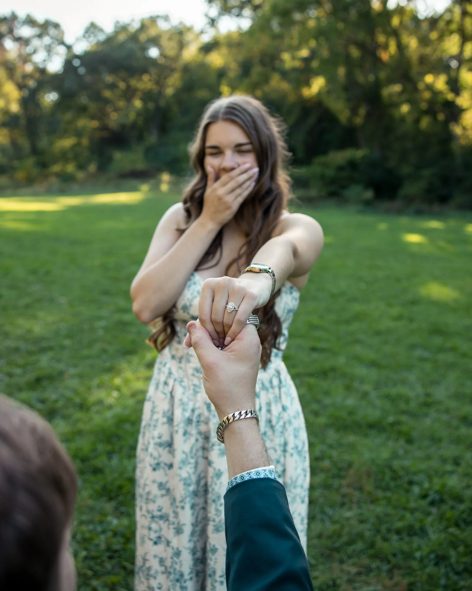 A woman in a floral dress is surprised and happy as a man shows her his engagement ring, outdoors in a park with green grass and trees.