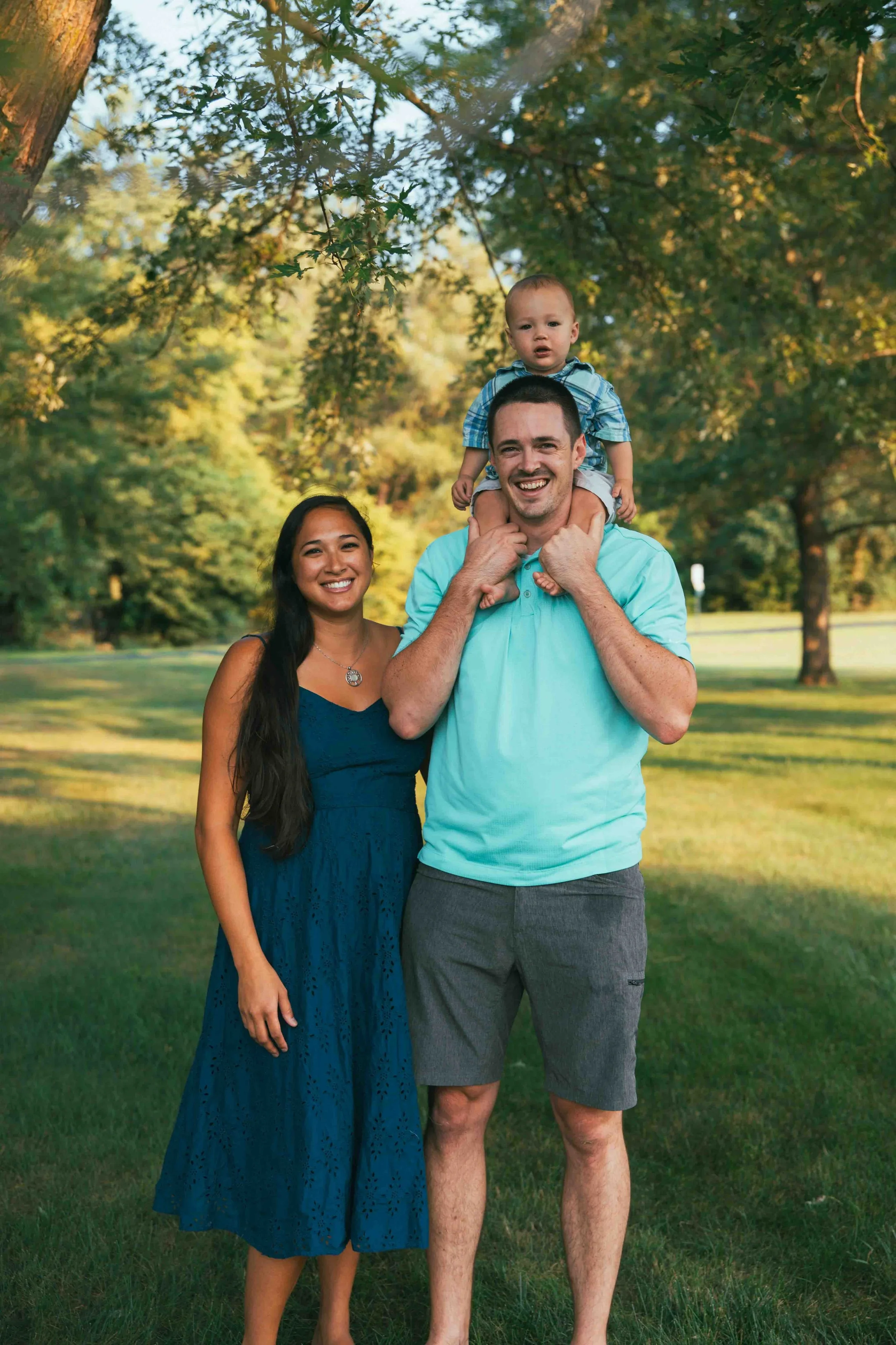 Smiling family of three standing outside in a park during daytime, with the father carrying a young child on his shoulders and the mother standing next to them.