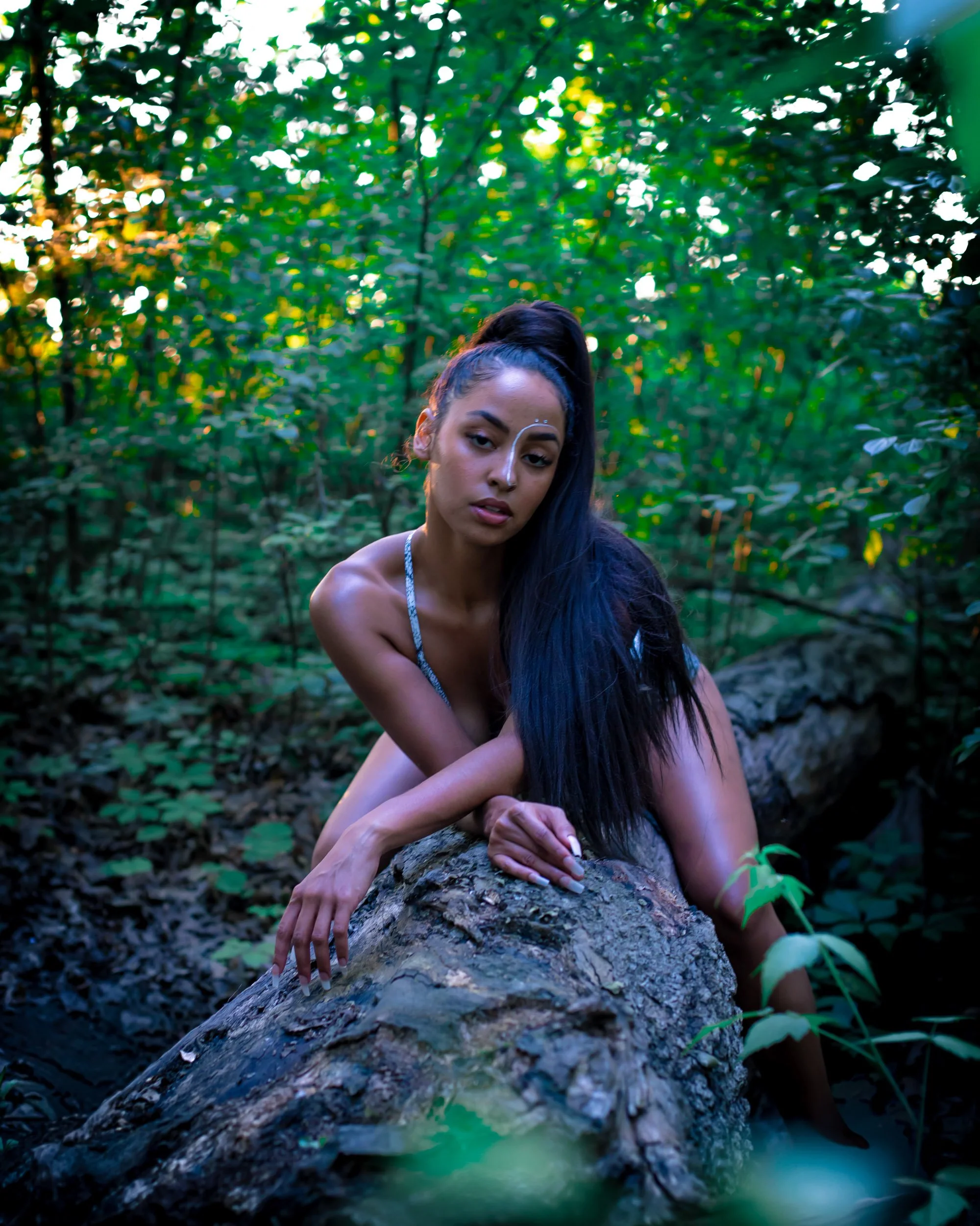 A woman with long black hair and light brown skin in a forest, leaning on a fallen tree trunk, looking at the camera with a contemplative expression.