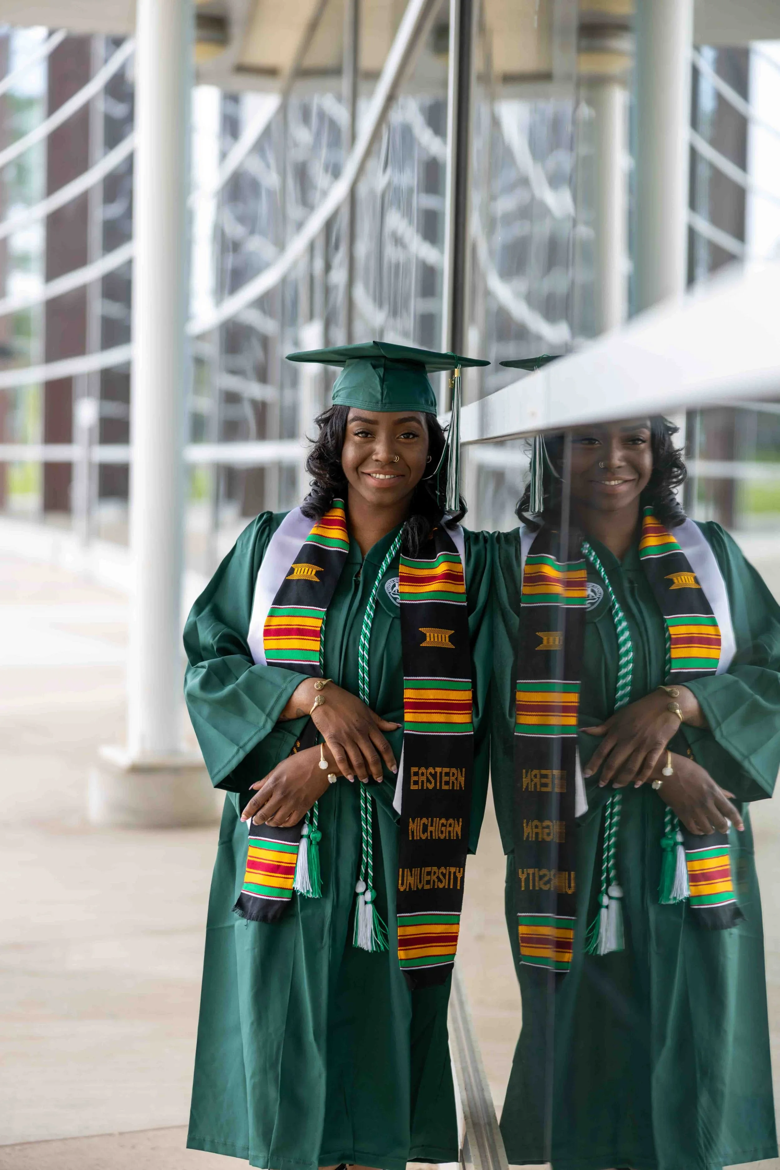 A young woman in a green graduation gown and cap, wearing a colorful Kente stole, stands beside a glass wall and looks at her reflection, smiling.