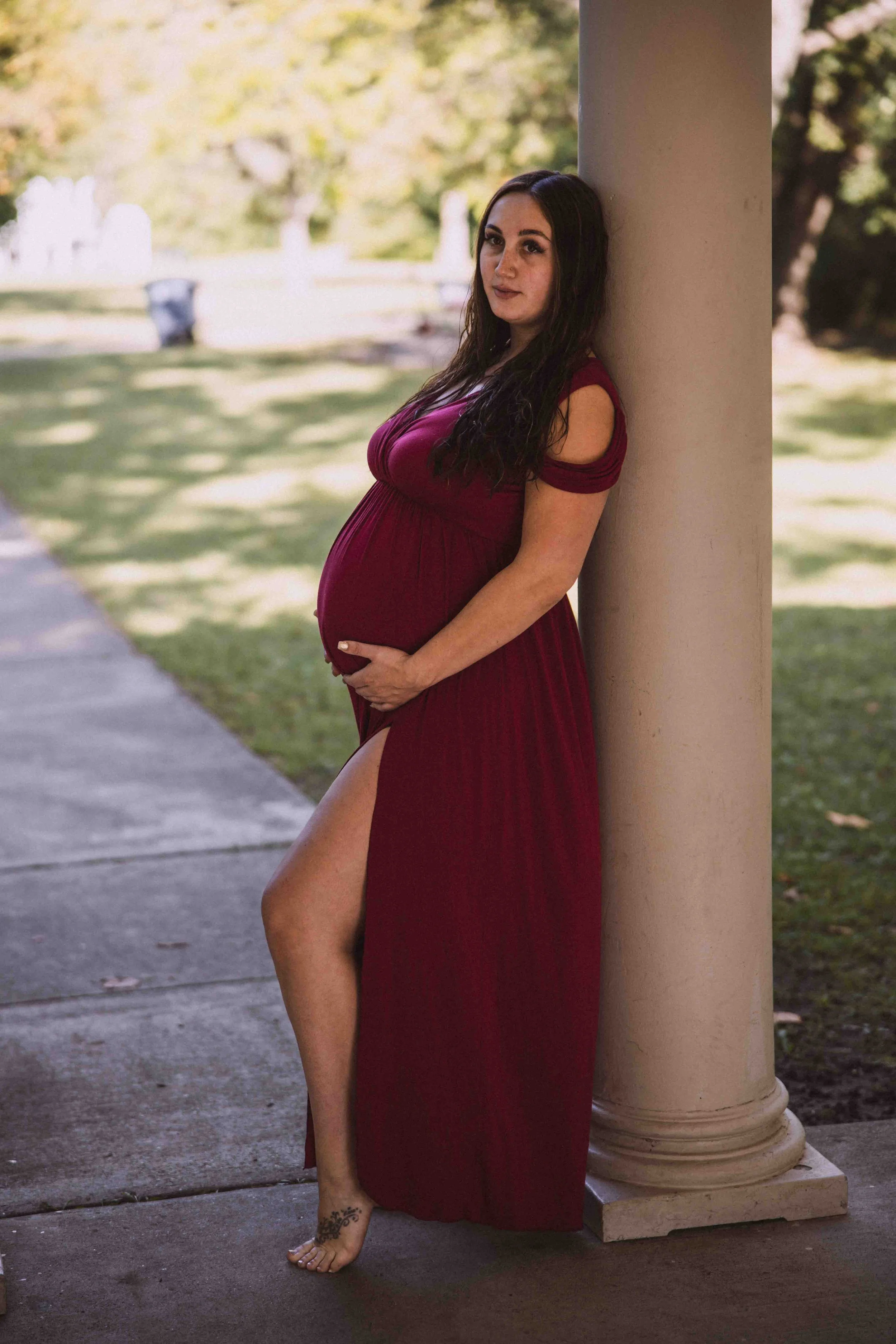 Pregnant woman in a long burgundy dress with a slit, posing outdoors next to a beige column, touching her belly, with green trees and a sidewalk in the background.