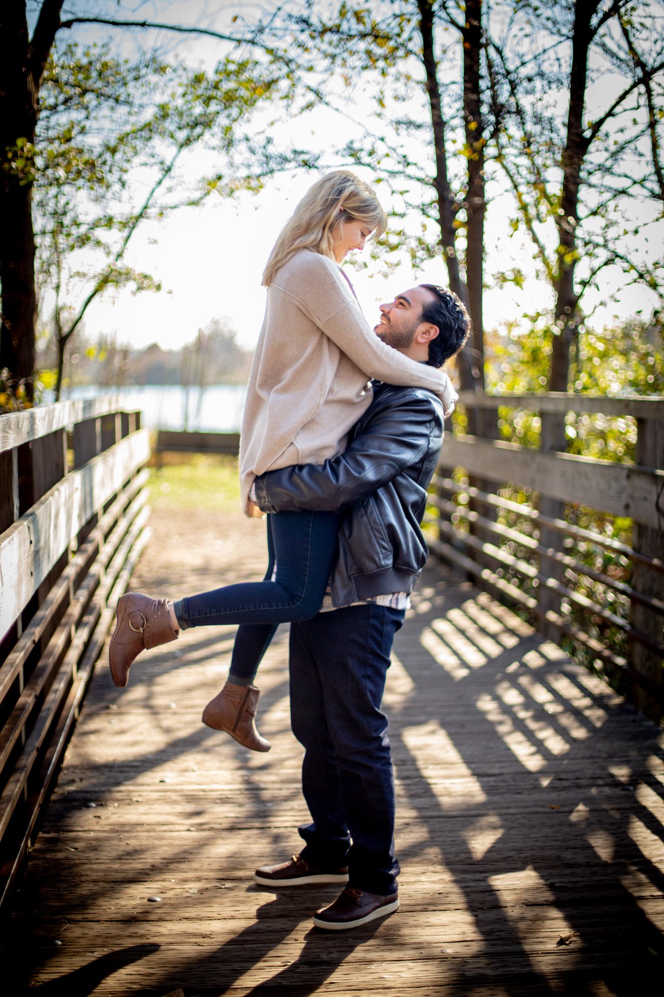 Westland Michigan engagement photography of A man with dark hair and a beard lifting a woman with blonde hair on a wooden bridge outdoors, with trees and sunlight in the background.