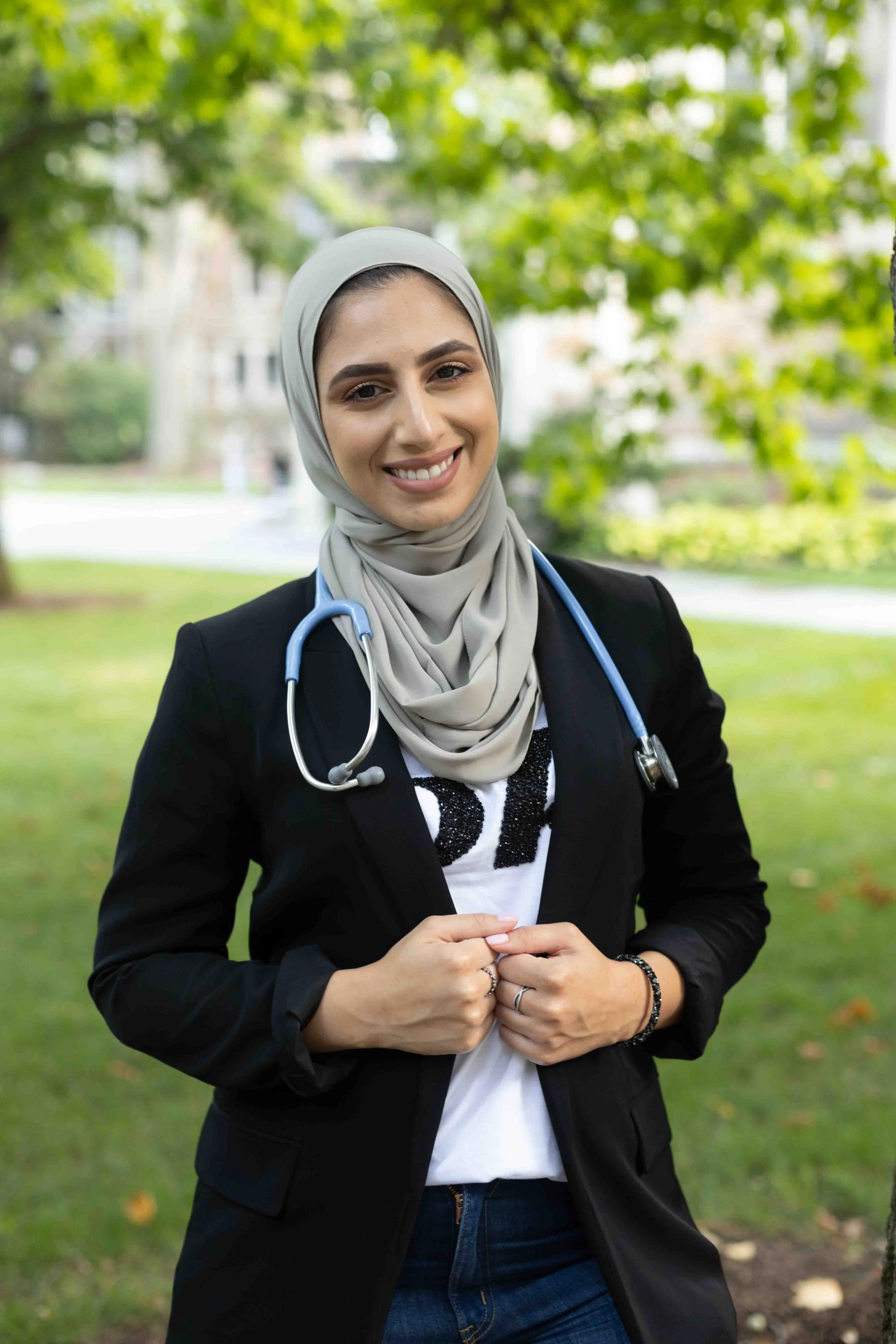 A young woman wearing a hijab, blazer, and stethoscope, standing outdoors in a park with trees and grass, smiling at the camera.
