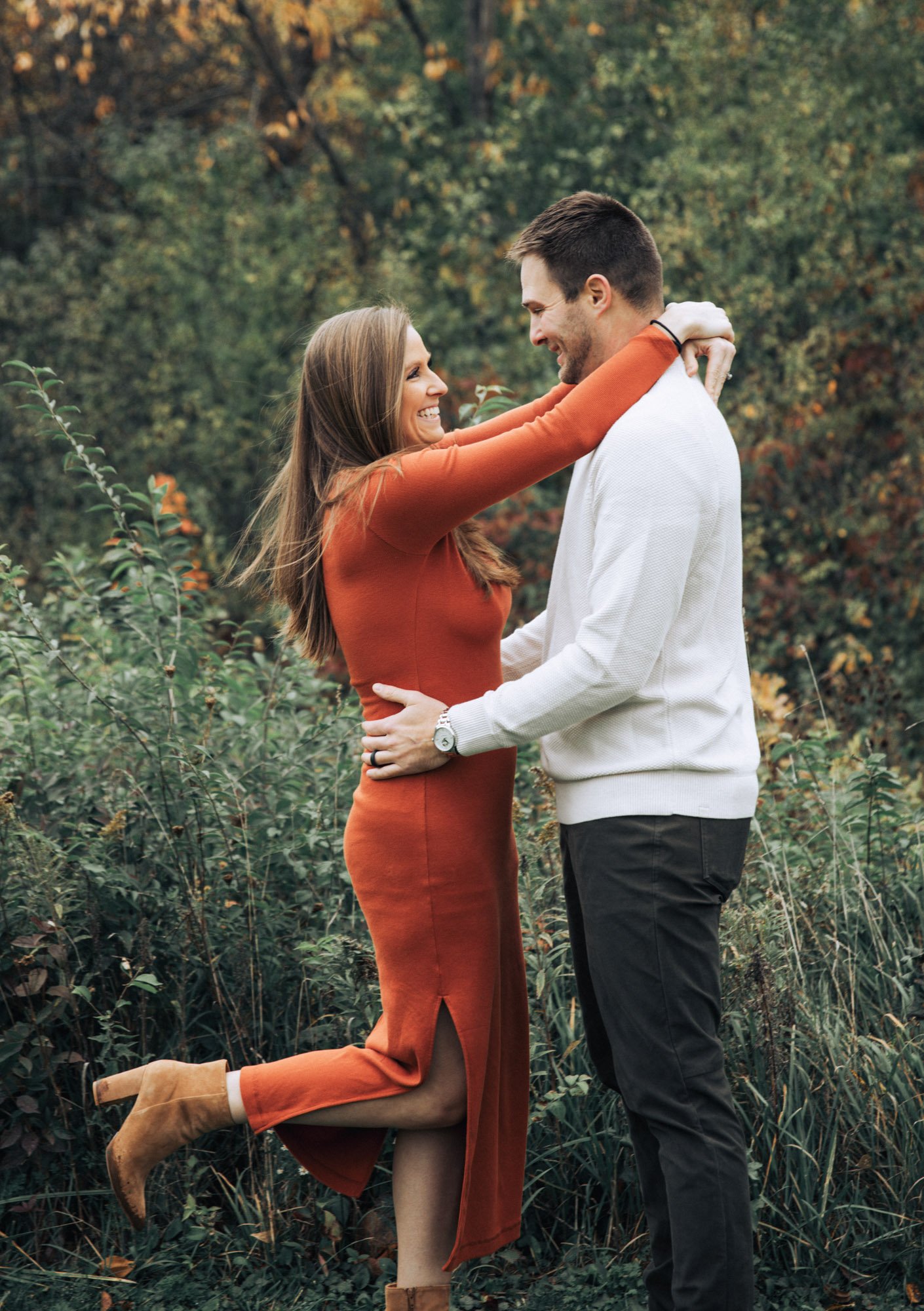 A smiling couple embracing outdoors among trees. The woman, in an orange dress, is lifting her leg and wrapping her arms around the man's neck. The man, in a white sweater and black pants, holds her waist. They appear happy and affectionate.