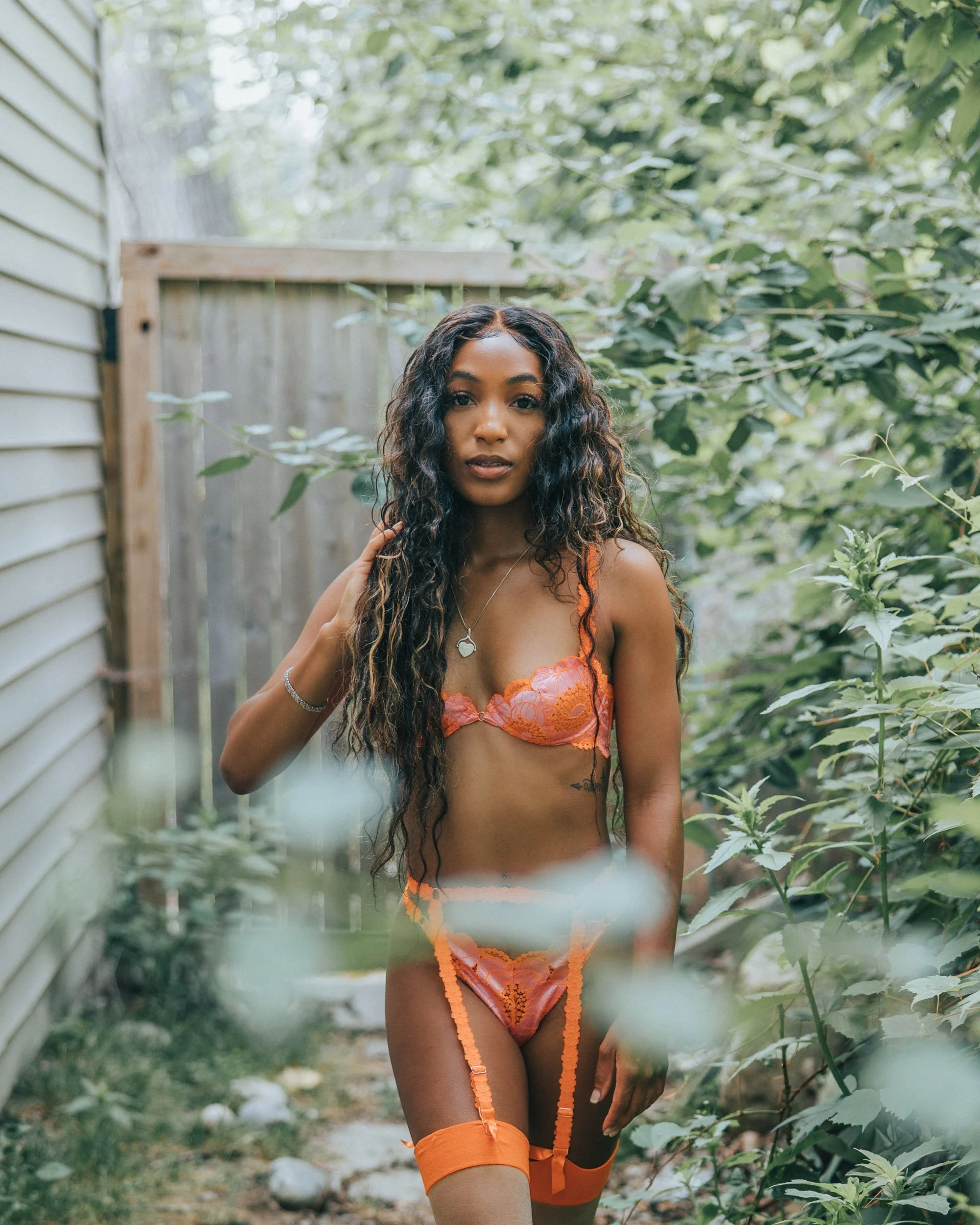 A woman in orange lingerie standing in a lush green garden next to a wooden fence.