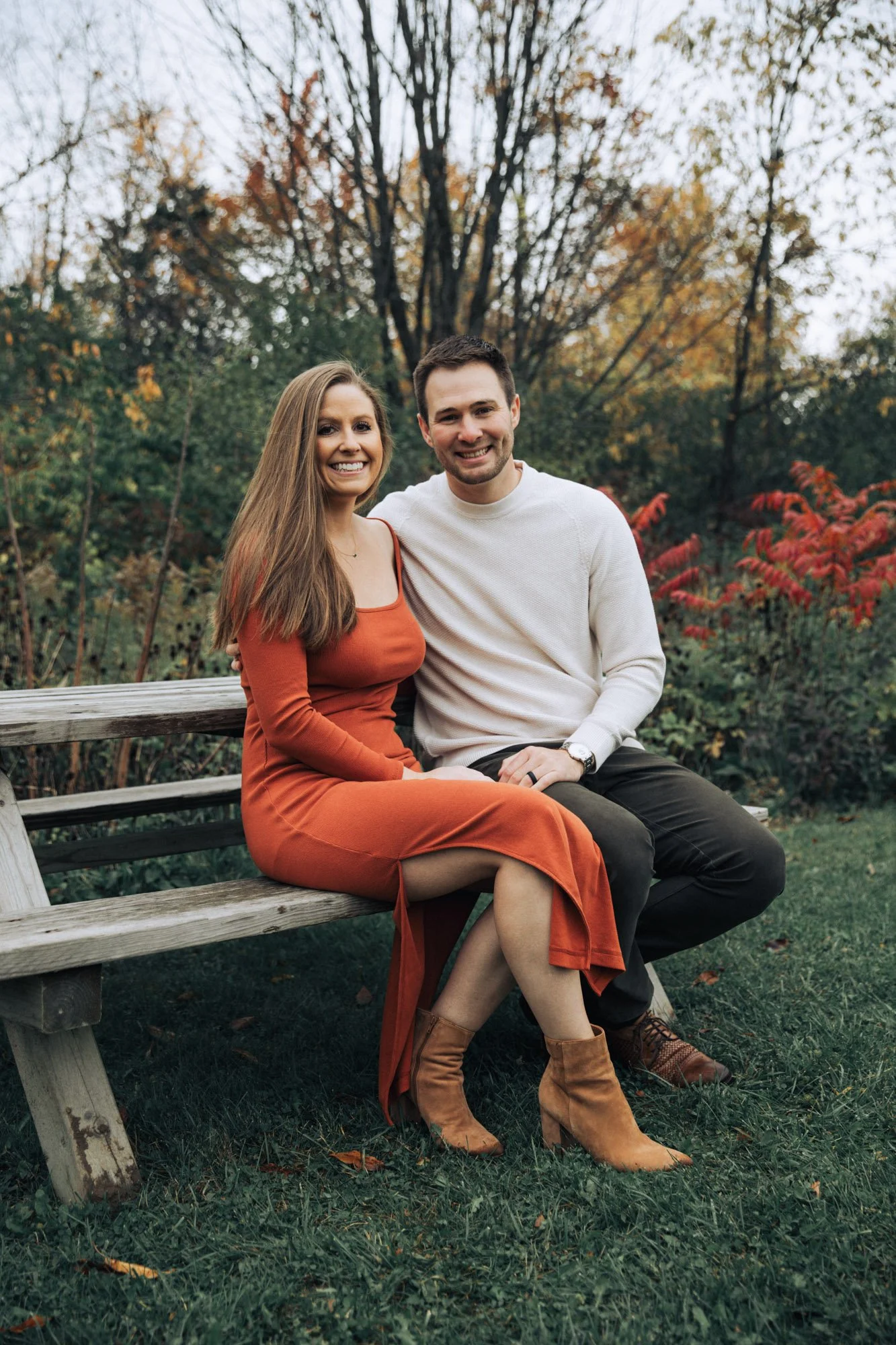 A smiling couple sitting on a wooden park bench during fall, surrounded by colorful autumn trees and grass.