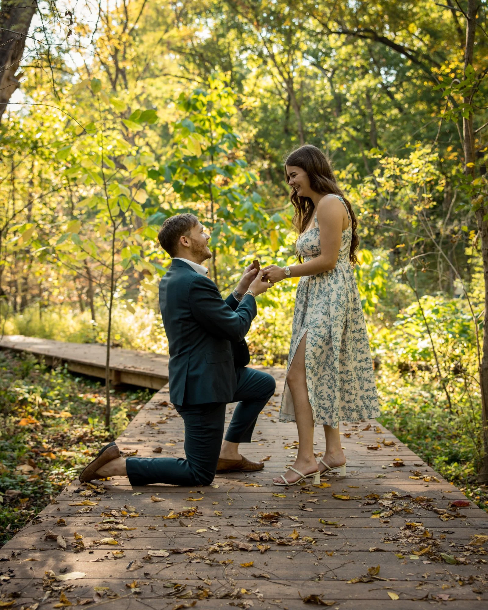 A man in a suit on one knee proposing to a woman in a floral dress on a wooden pathway in a forest.