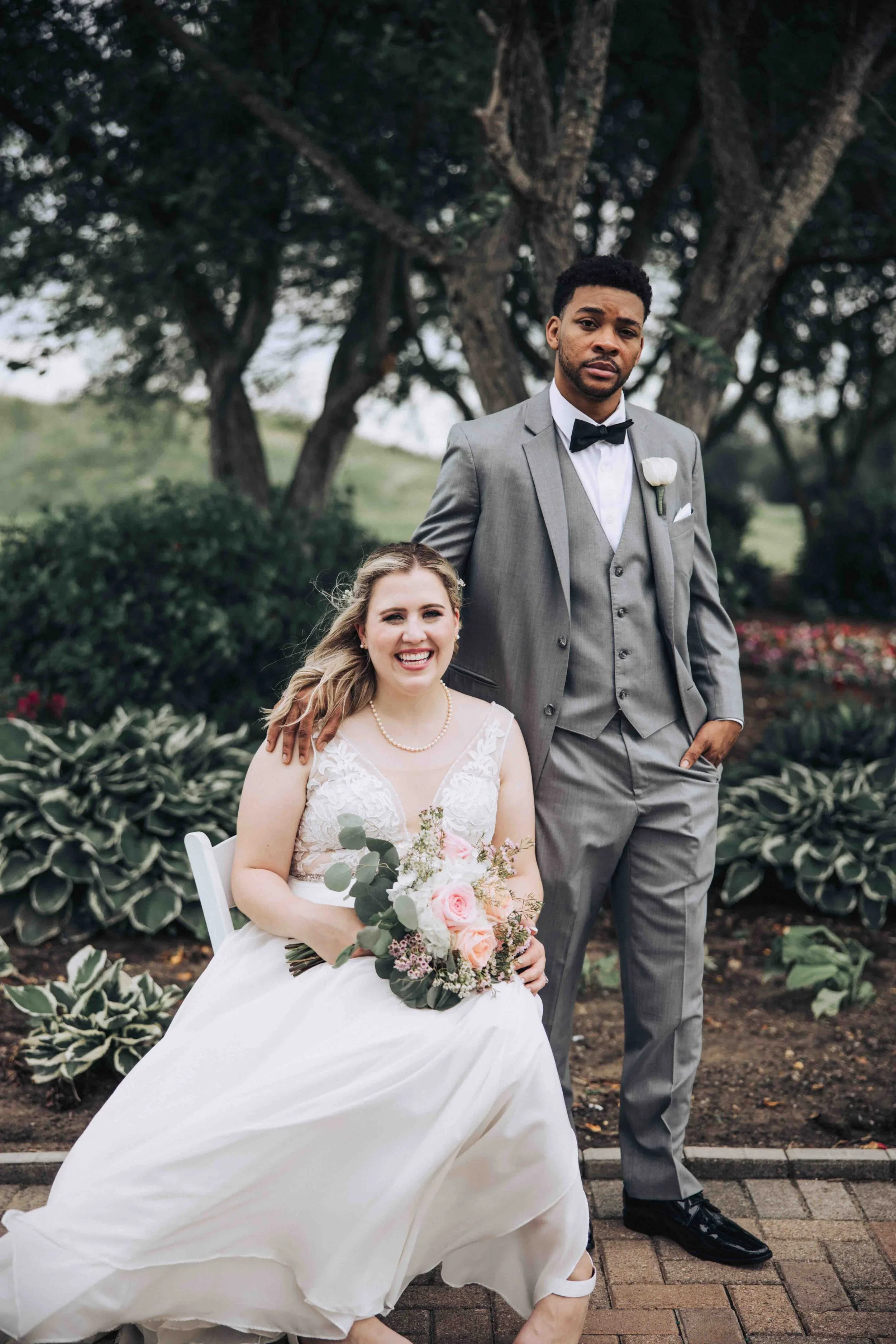 Bride in a white wedding dress sitting with a bouquet of pink and white flowers, groom in a gray suit with a bow tie standing beside her outdoors.
