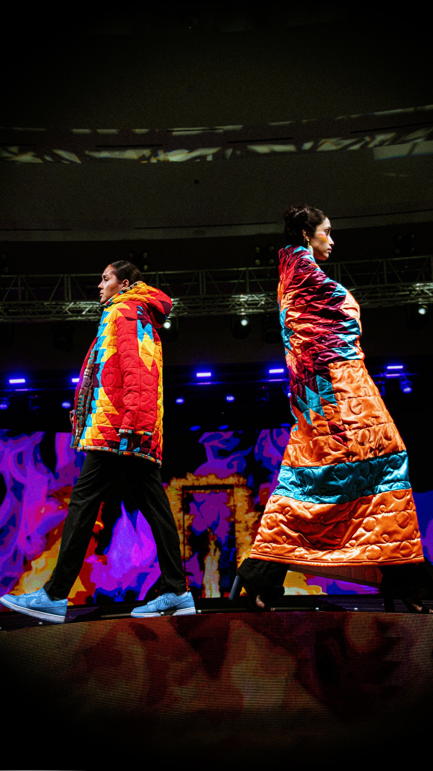 Two models walking on a runway wearing colorful, quilted, patterned coats in a fashion show with bright stage lights and digital screens in the background.