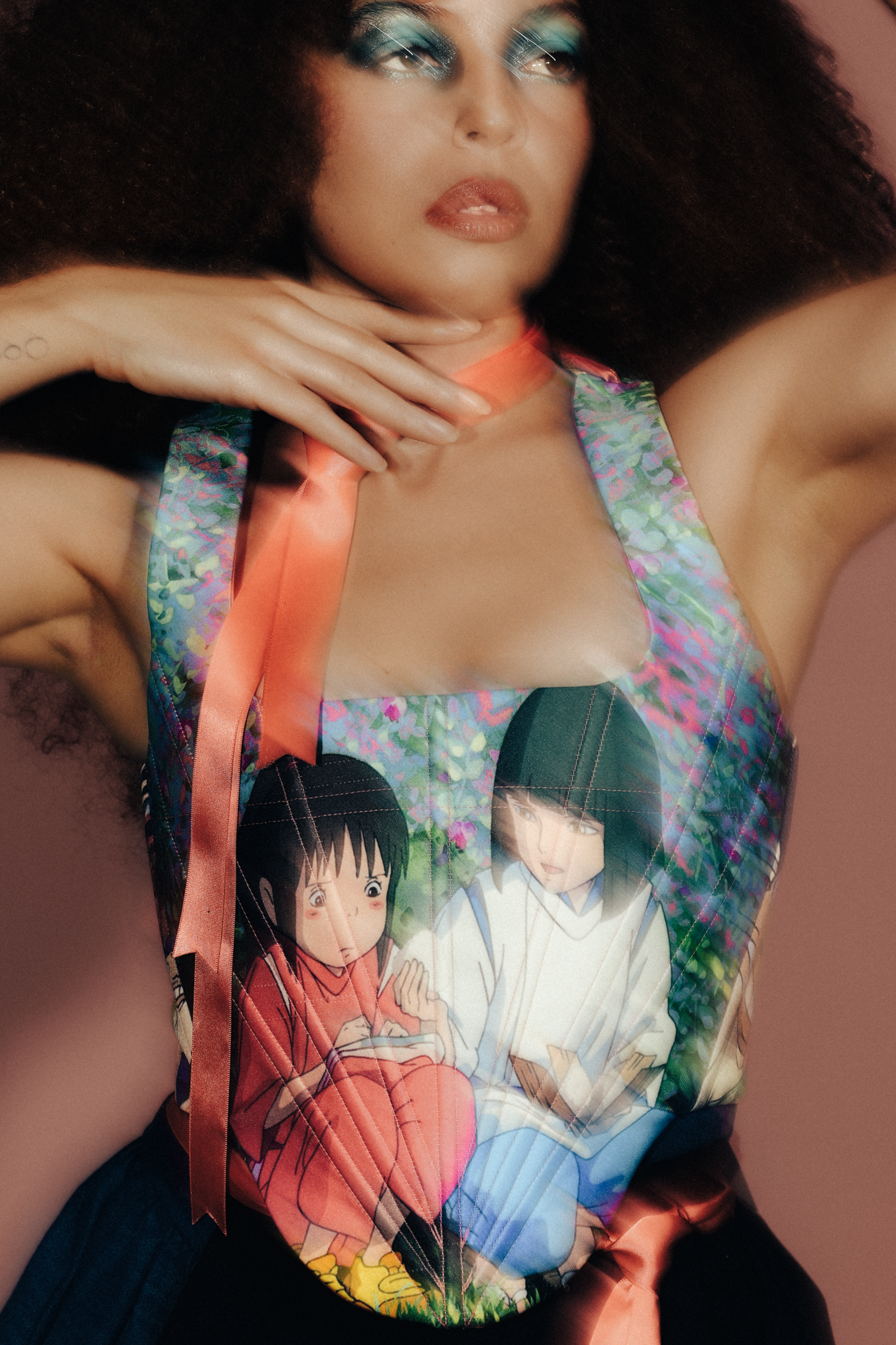 A woman with curly hair and bold blue eye makeup poses with her arms raised, wearing a sleeveless top, or corset, featuring characters from an anime show called Spirited Away, against a pink background, photographed during a fashion editorial photogr