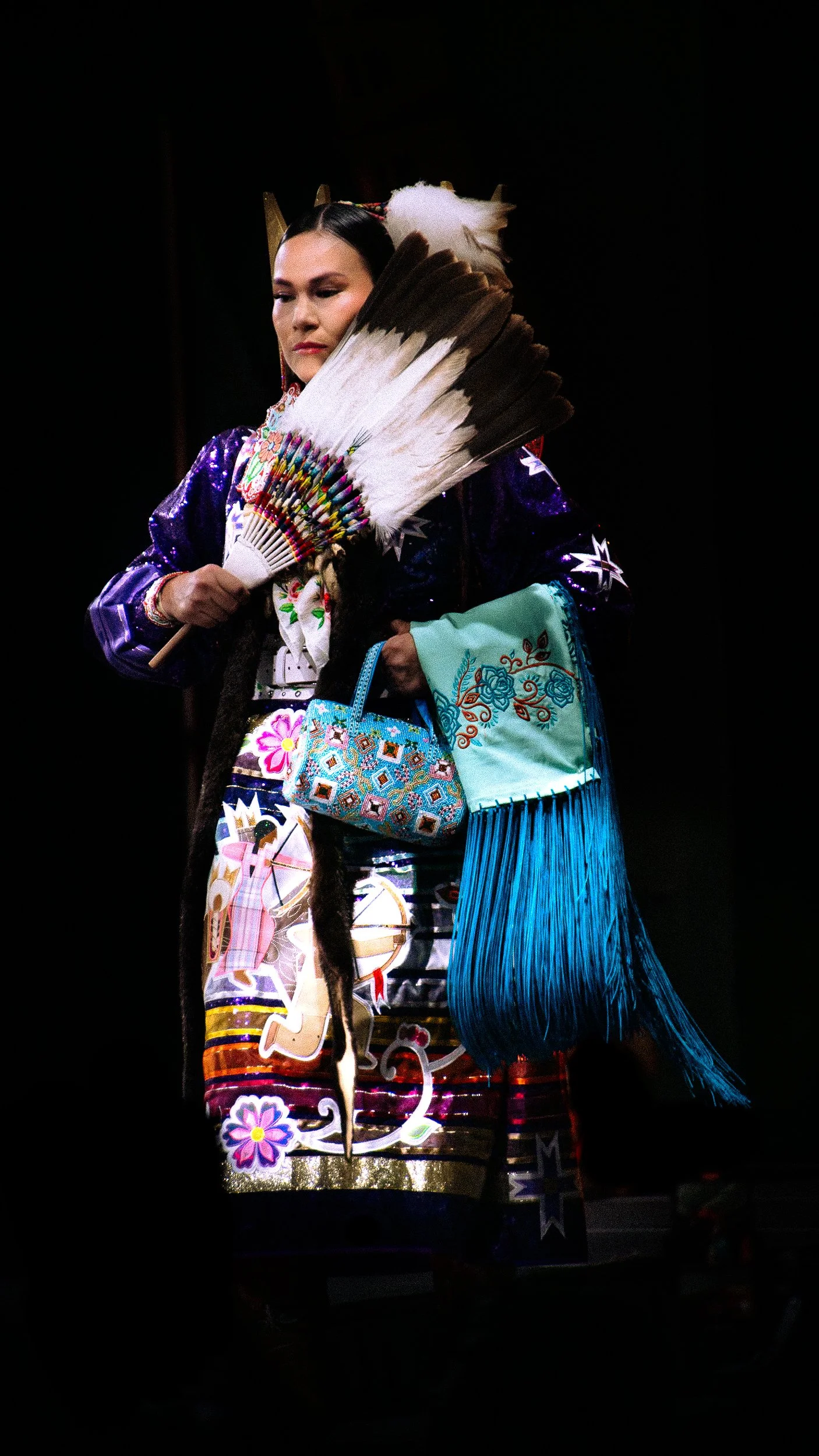 Woman in traditional indigenous Aztec clothing holding a feathered fan, carrying embroidered bags on a dark background.