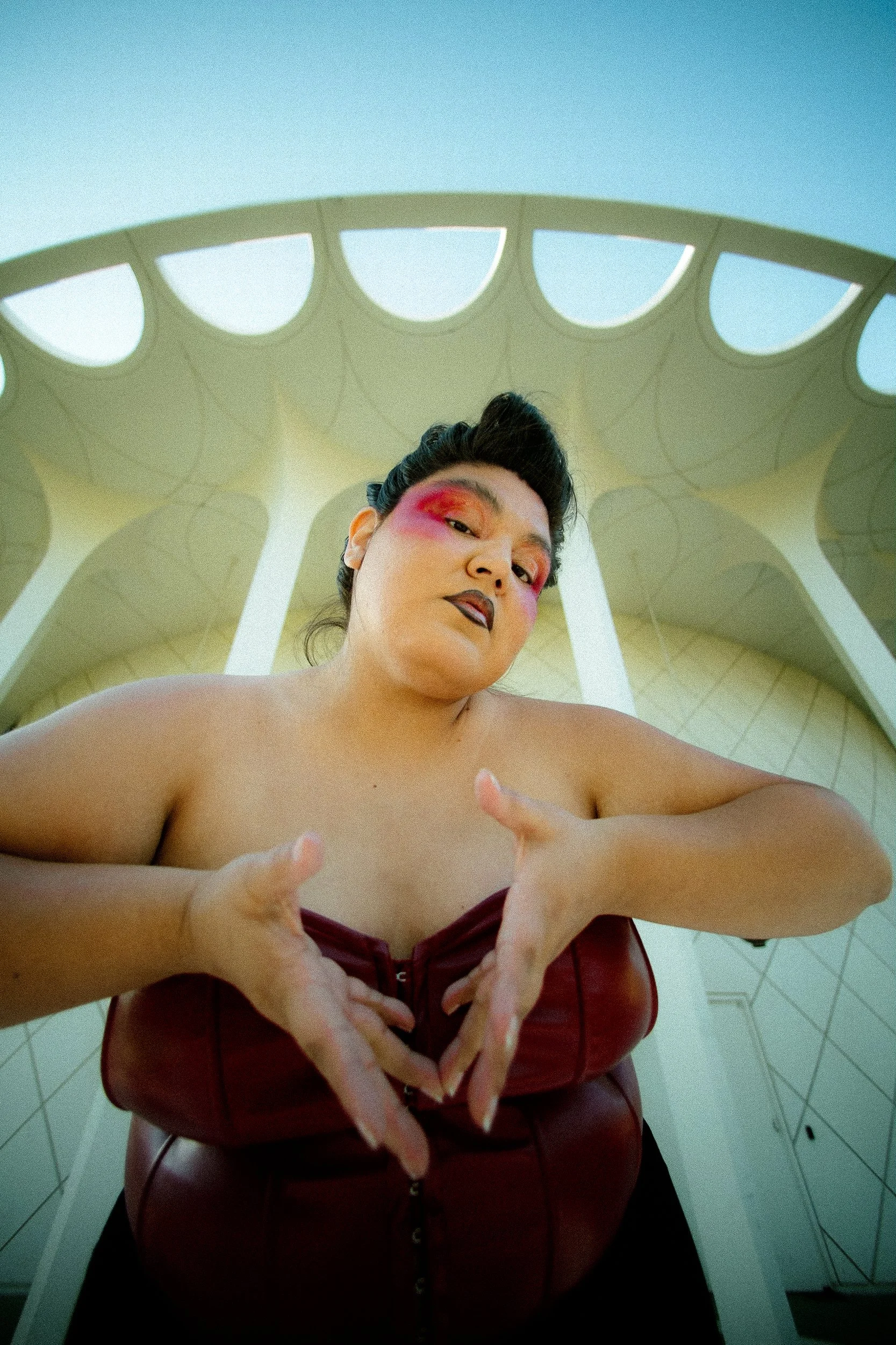 A Latina woman model with bold makeup, wearing a red leather top, standing outdoors under a modern architectural structure with a sky background, photographed during a fashion editorial photography session in Los Angeles with fashion photographer Gí 