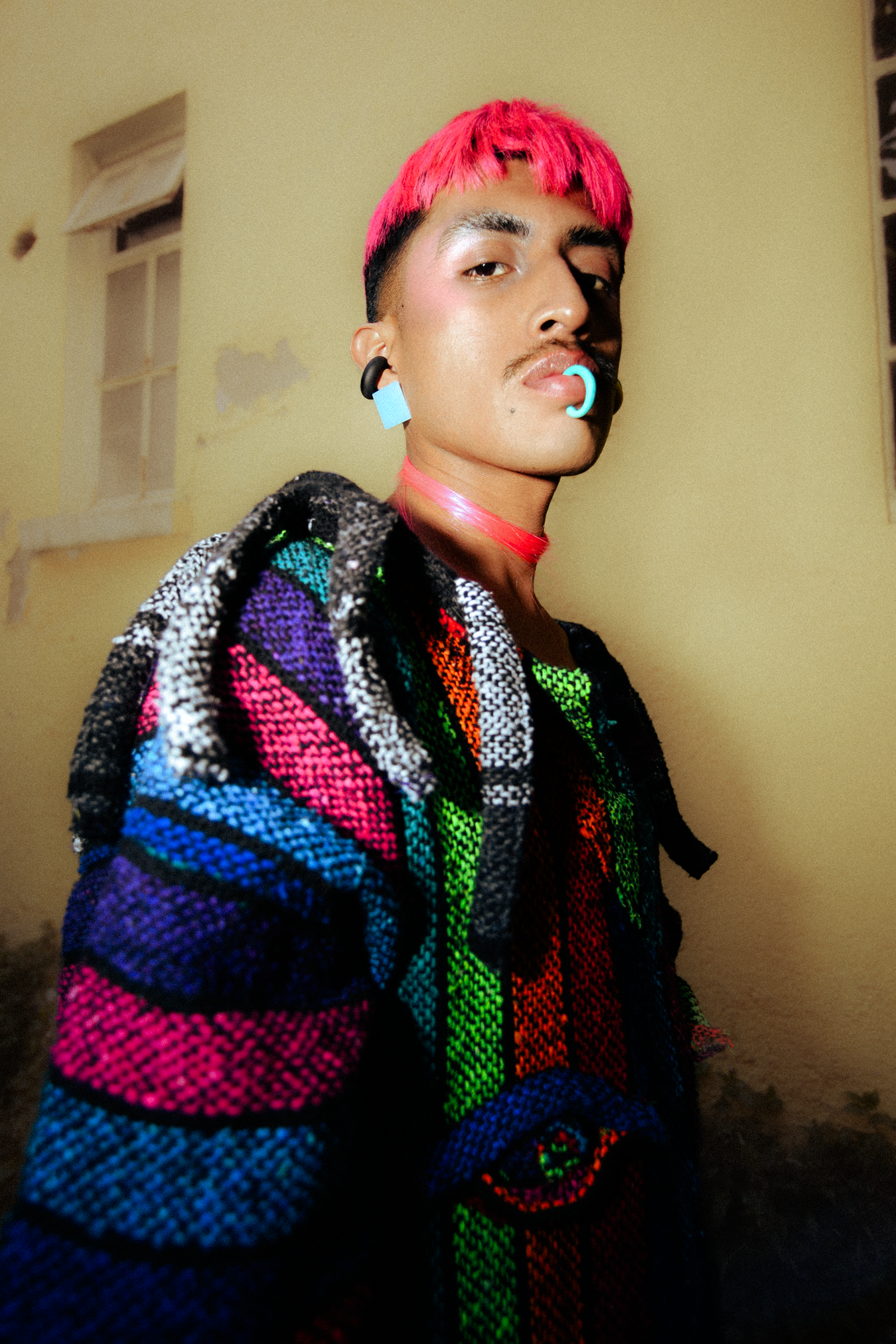 A brown person with pink hair and colorful Pancho-inspired piece standing in a garden outdoor area, shot for an editorial fashion photography session in Los Angeles with fashion photographer Gí Tenamá..