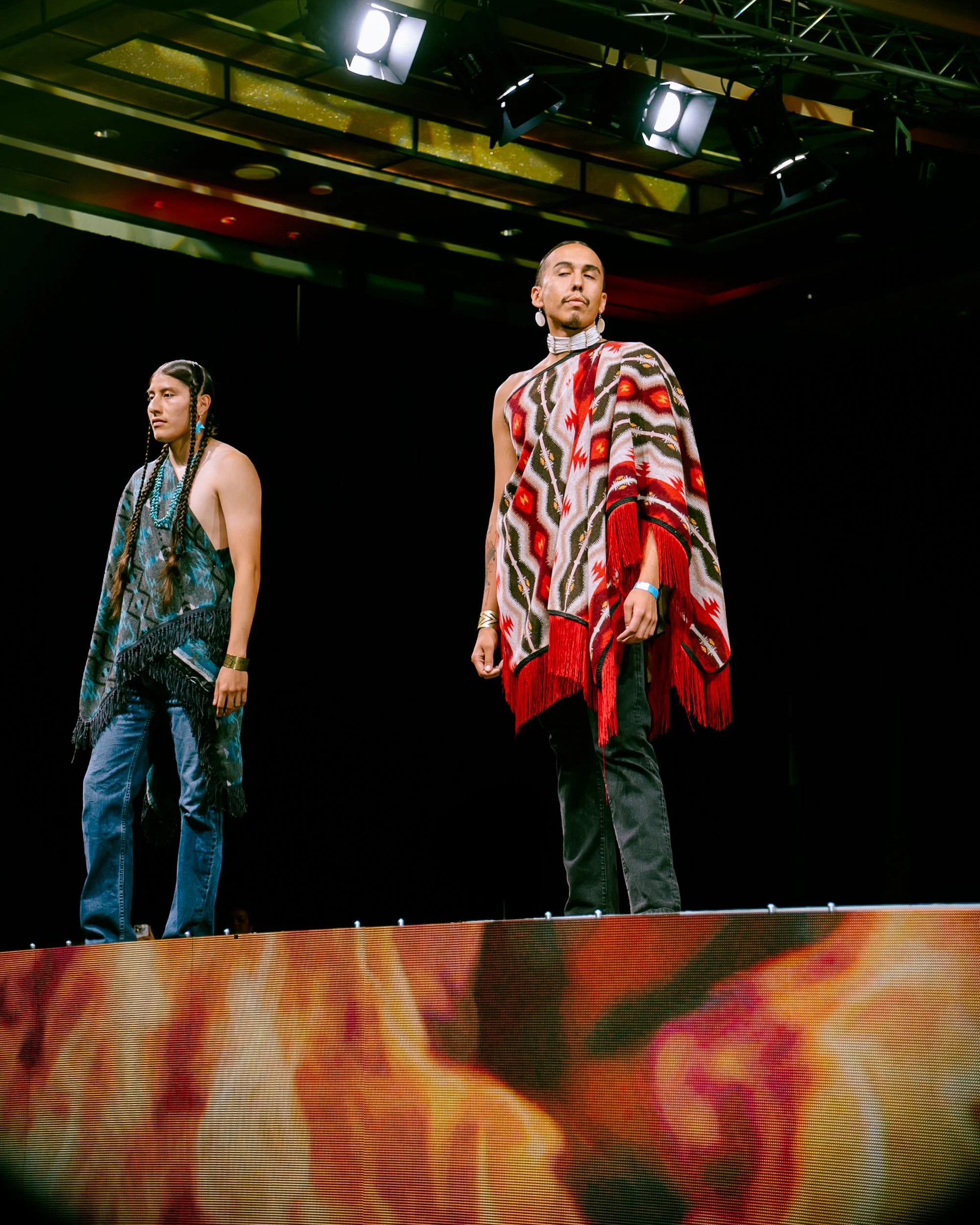 Two models on a runway stage wearing colorful, patterned fashion outfits, with one wearing a fringed shawl and the other with braided hair, under bright overhead stage lights.