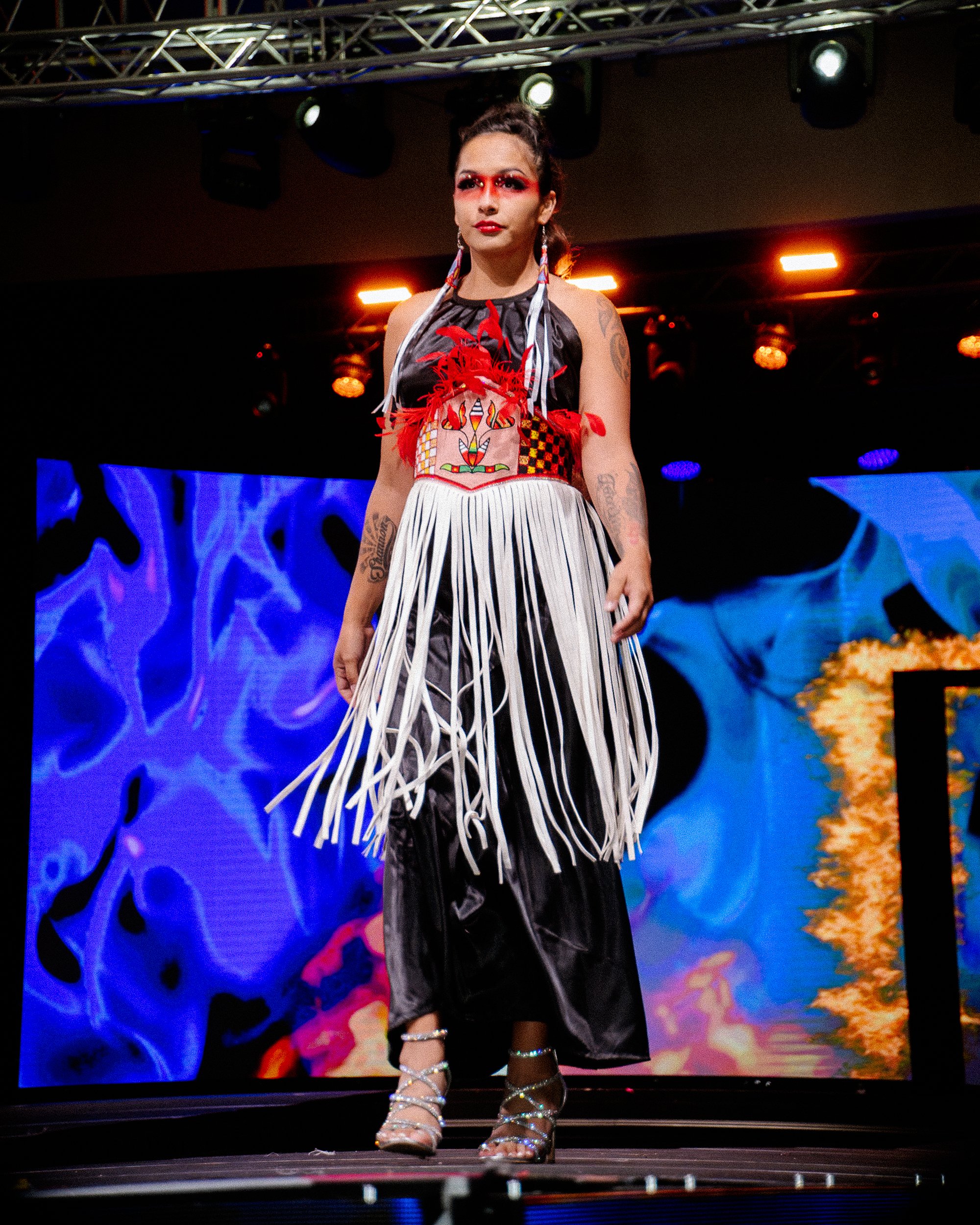 A woman walking on a runway during a fashion show, wearing a colorful, artistic dress with fringes and metallic heels.