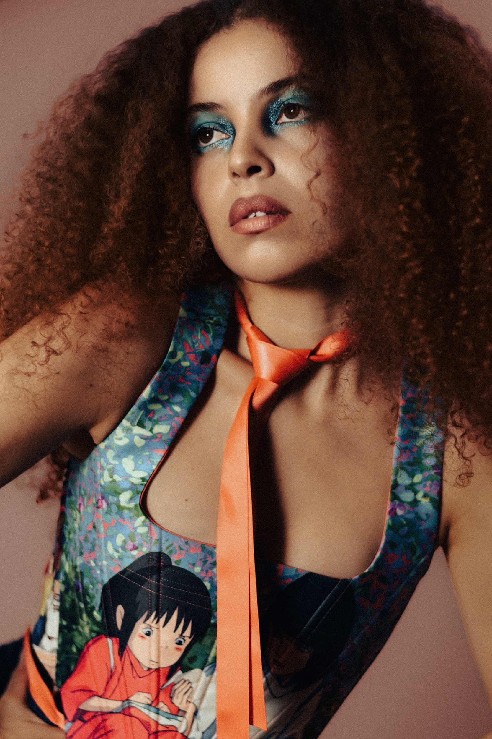 A woman with curly hair and bold blue eye makeup, wearing a dress with an anime character print and an orange necktie, against a plain background, photographed during a fashion editorial photography session in Los Angeles with fashion photographer Gí