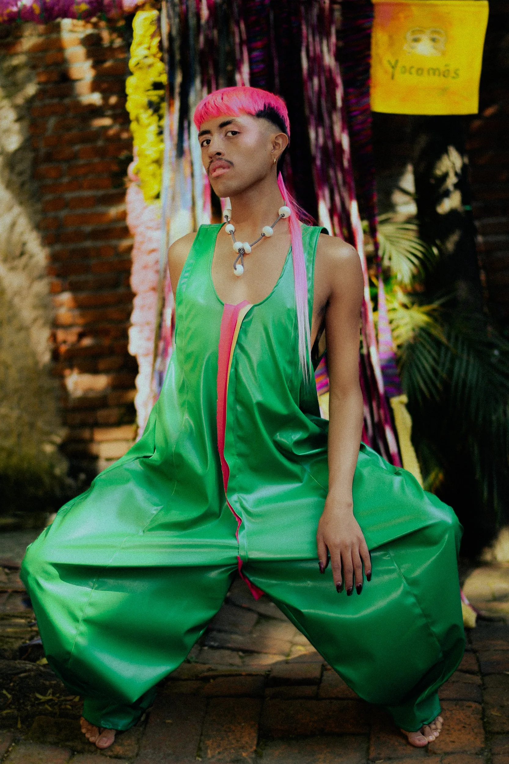 A feminine model  with pink hair and mustache, wearing green baggy jumpsuit, sitting on cobblestone ground, with colorful fabric backdrop and plants in background, photographed during a fashion editorial photography session in Los Angeles with fashio
