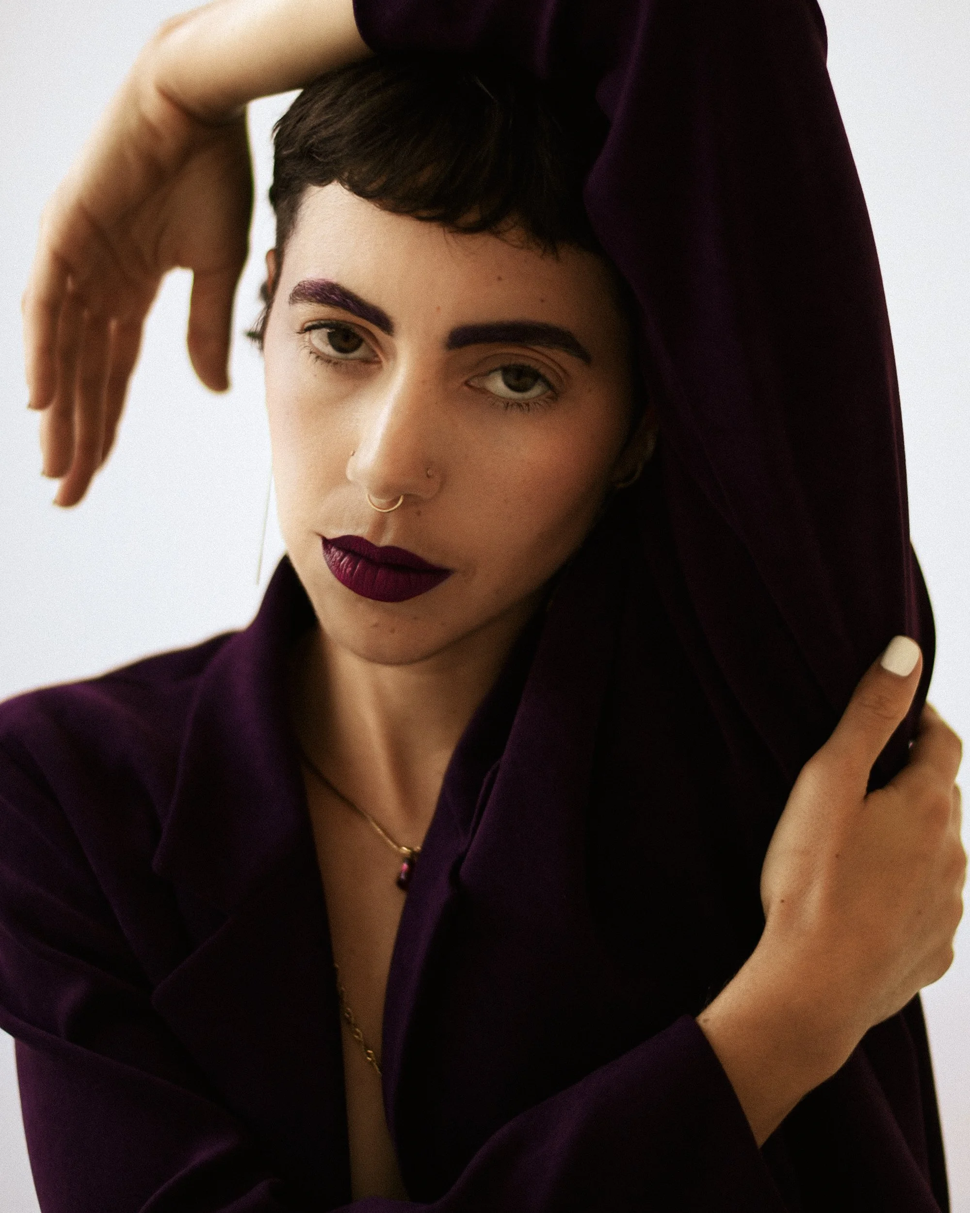 Close-up portrait of a queer nonbinary person with short, dark hair, purple eyebrows, and dark purple lipstick, wearing jewelry including earrings and necklaces, dressed in a dark purple shirt, against a plain white background, photographed during a 