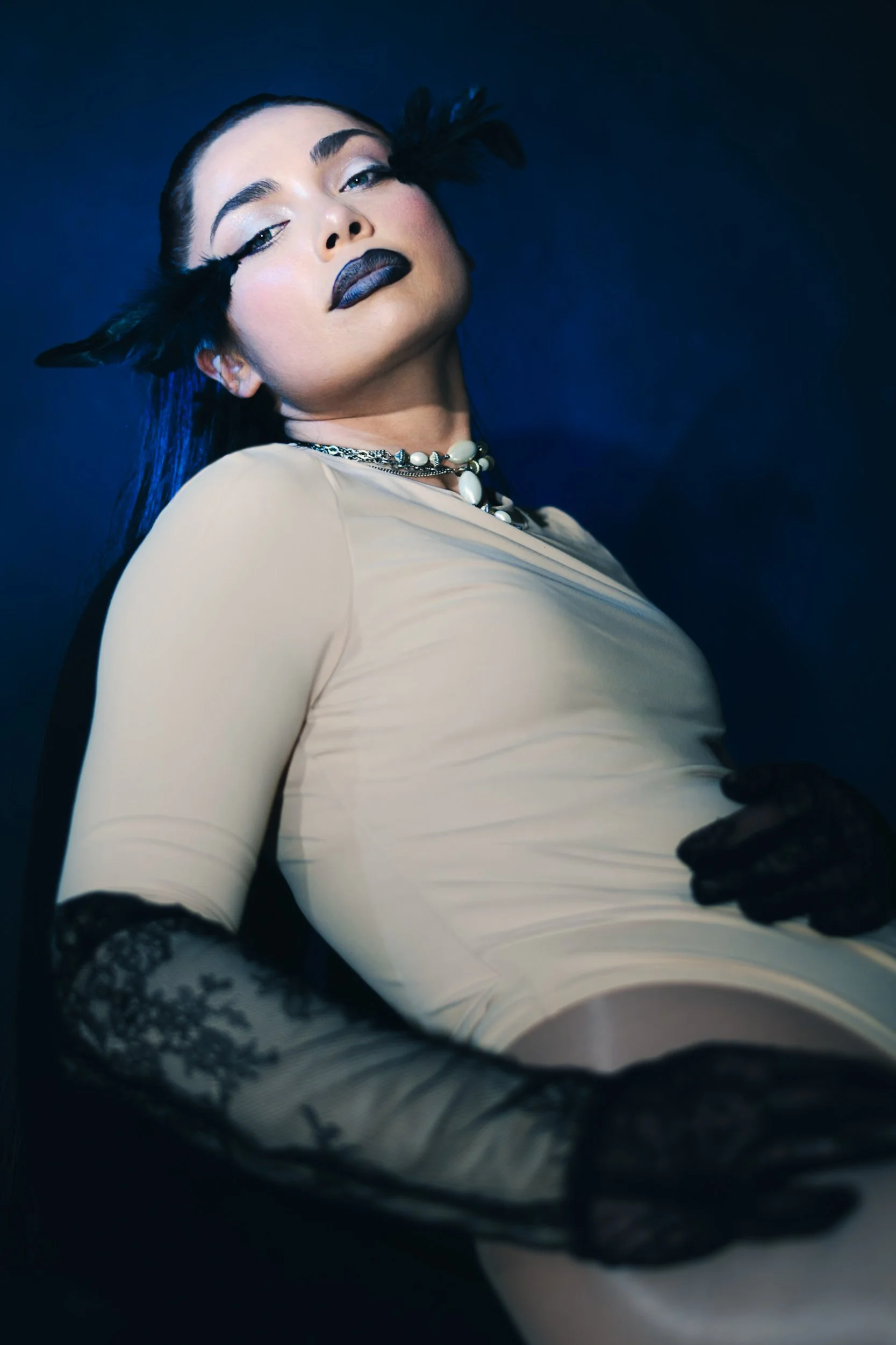 Southern California-based Indie artist and producer Doll Klaw poses in a studio in a beige leotard against a blue wall with black lace gloves, photo taken by Los Angeles photographer and director Gí Tenamá