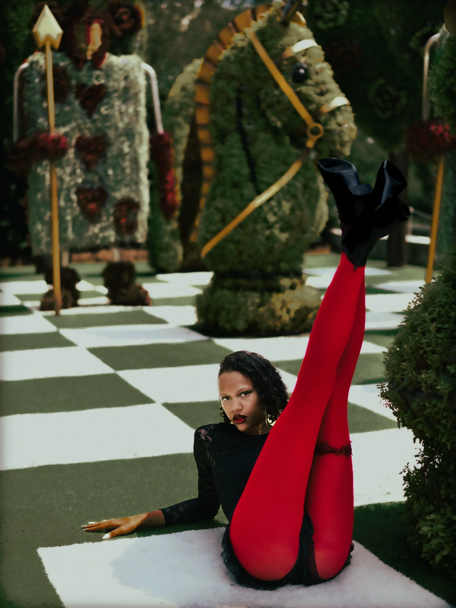 A woman model in black and red clothing poses on a black-and-white checkered floor with large decorative figures in the background, photographed during a fashion editorial photography session in Los Angeles with fashion photographer Gí Tenamá..
