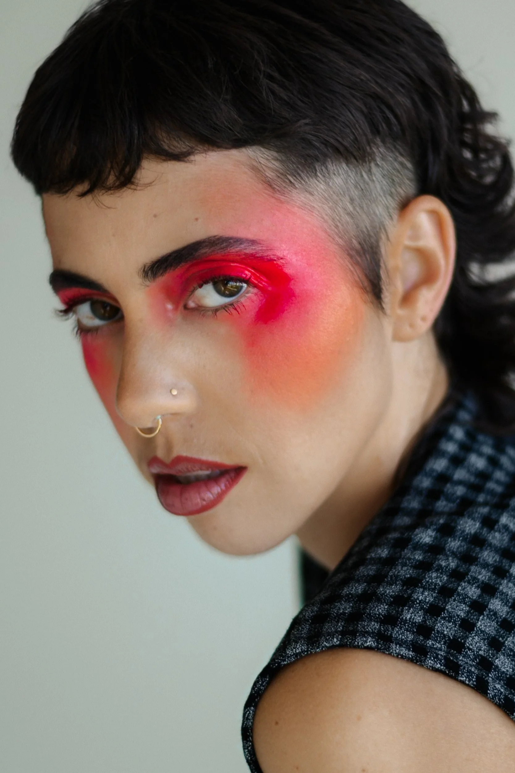 A person with short dark hair, green eyes, and multiple facial piercings wearing vibrant makeup with red and pink eyeshadow and lipstick, dressed in a black and white checkered top, photographed during a studio photography session in Los Angeles for 