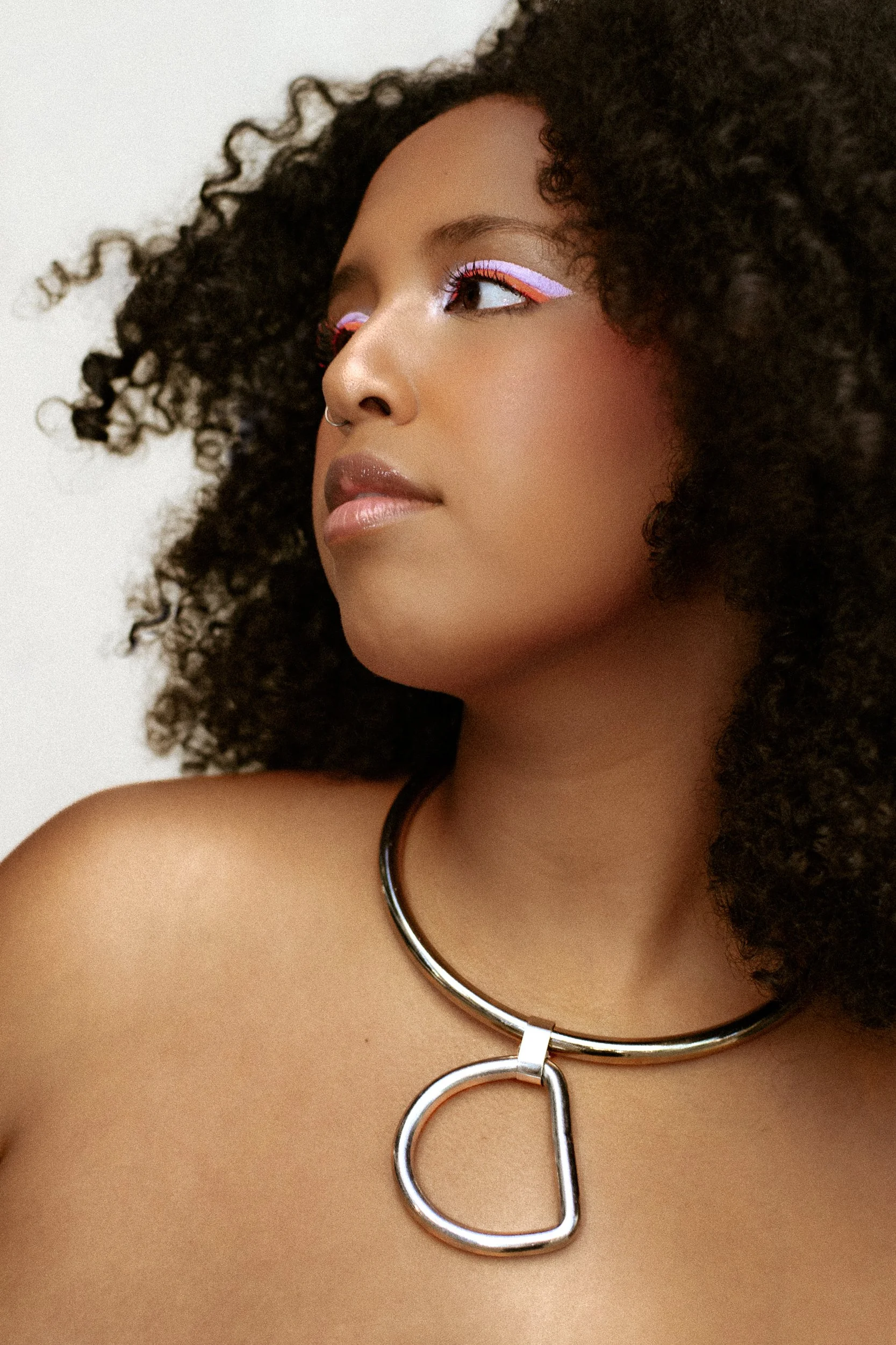 Close-up of a woman with curly hair, wearing colorful eye makeup and a silver necklace with a large D-shaped pendant, photographed during a studio photography session in Los Angeles for a fashion editorial collaboration with fashion photographer